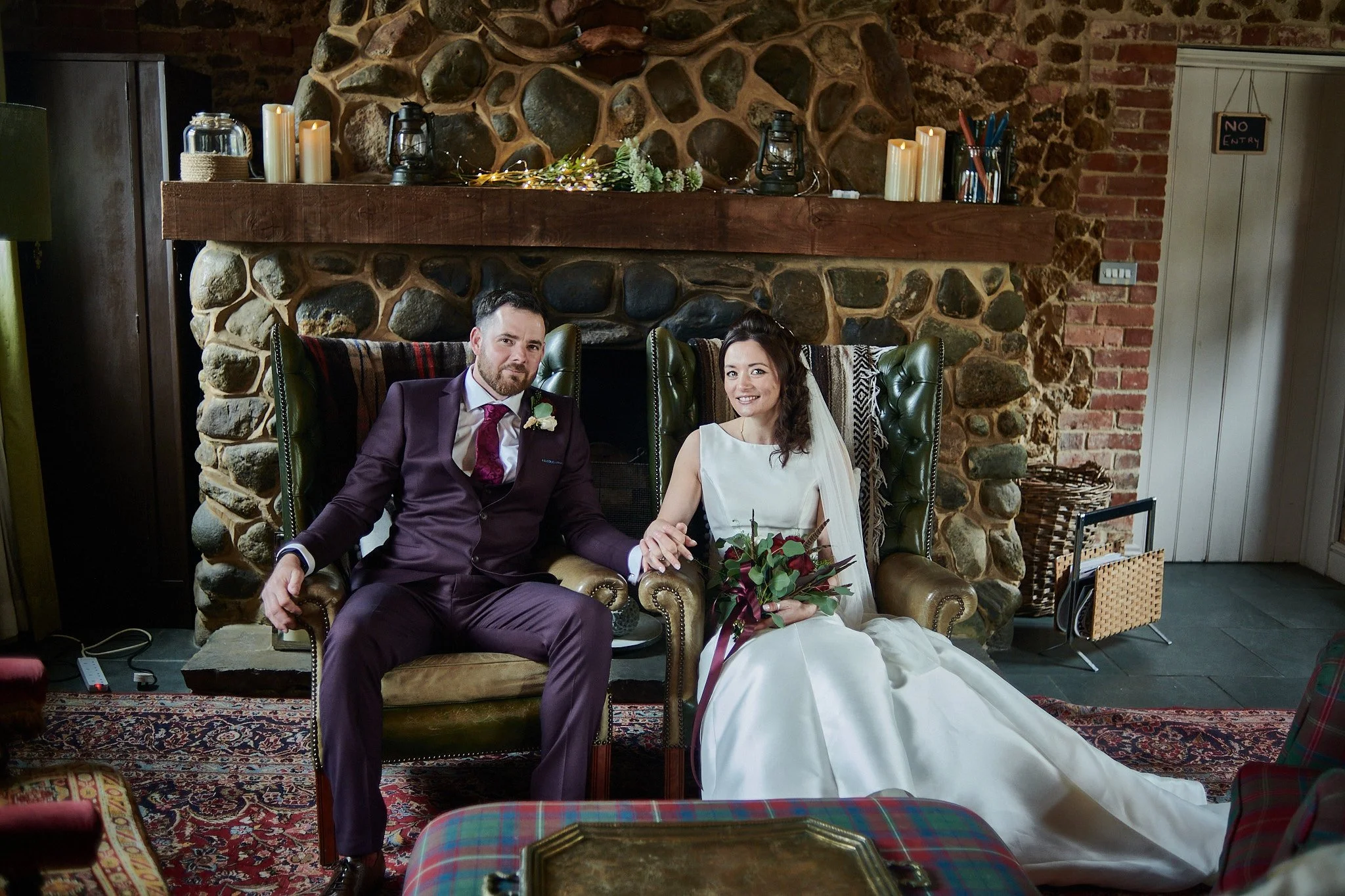 A bride and groom sitting together on a vintage green leather armchair in front of a stone fireplace, with candles and decorative items on the mantle.
