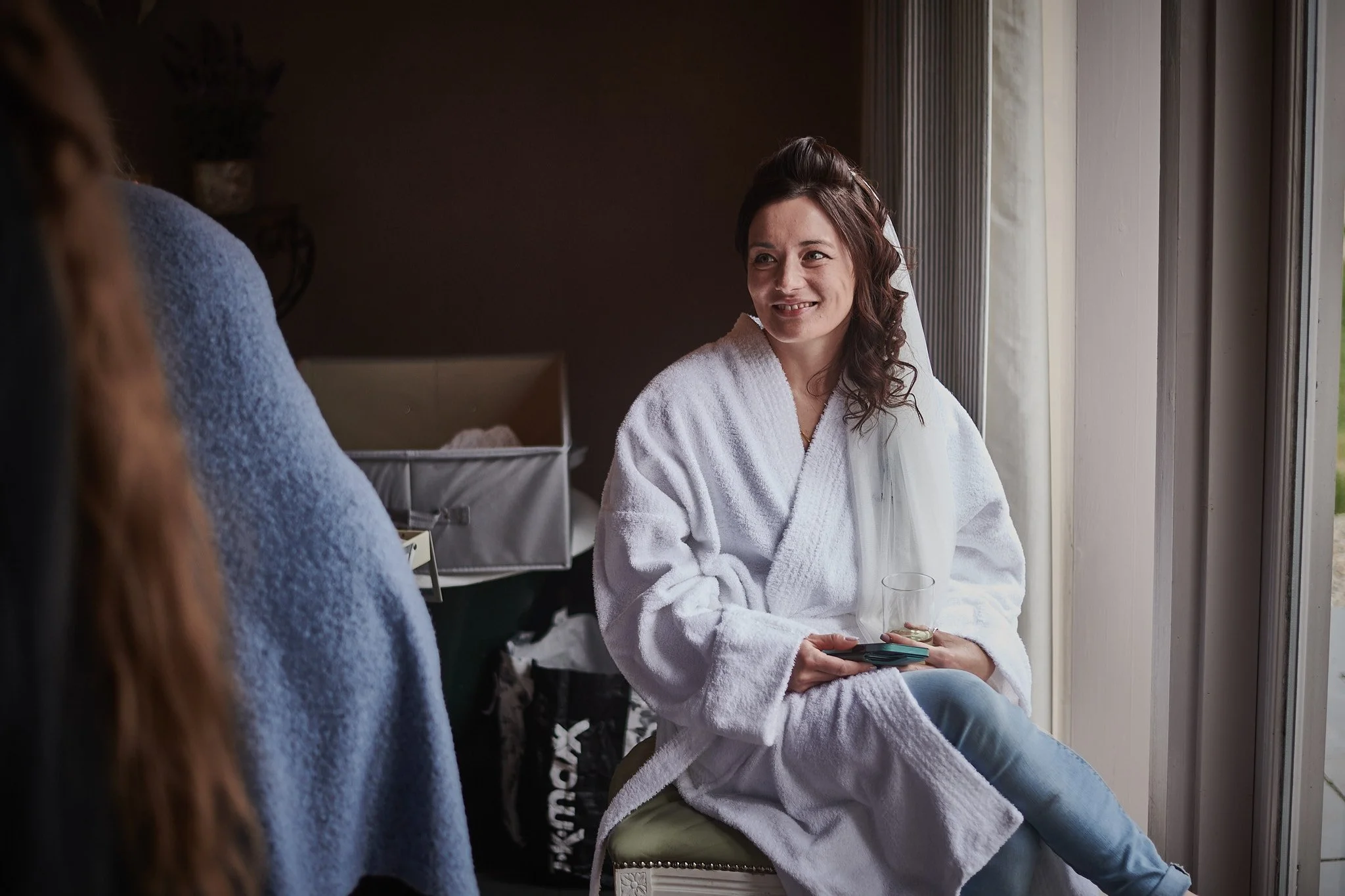 A woman in a white bathrobe sitting by a window, smiling, holding a glass, and talking to someone off-camera.