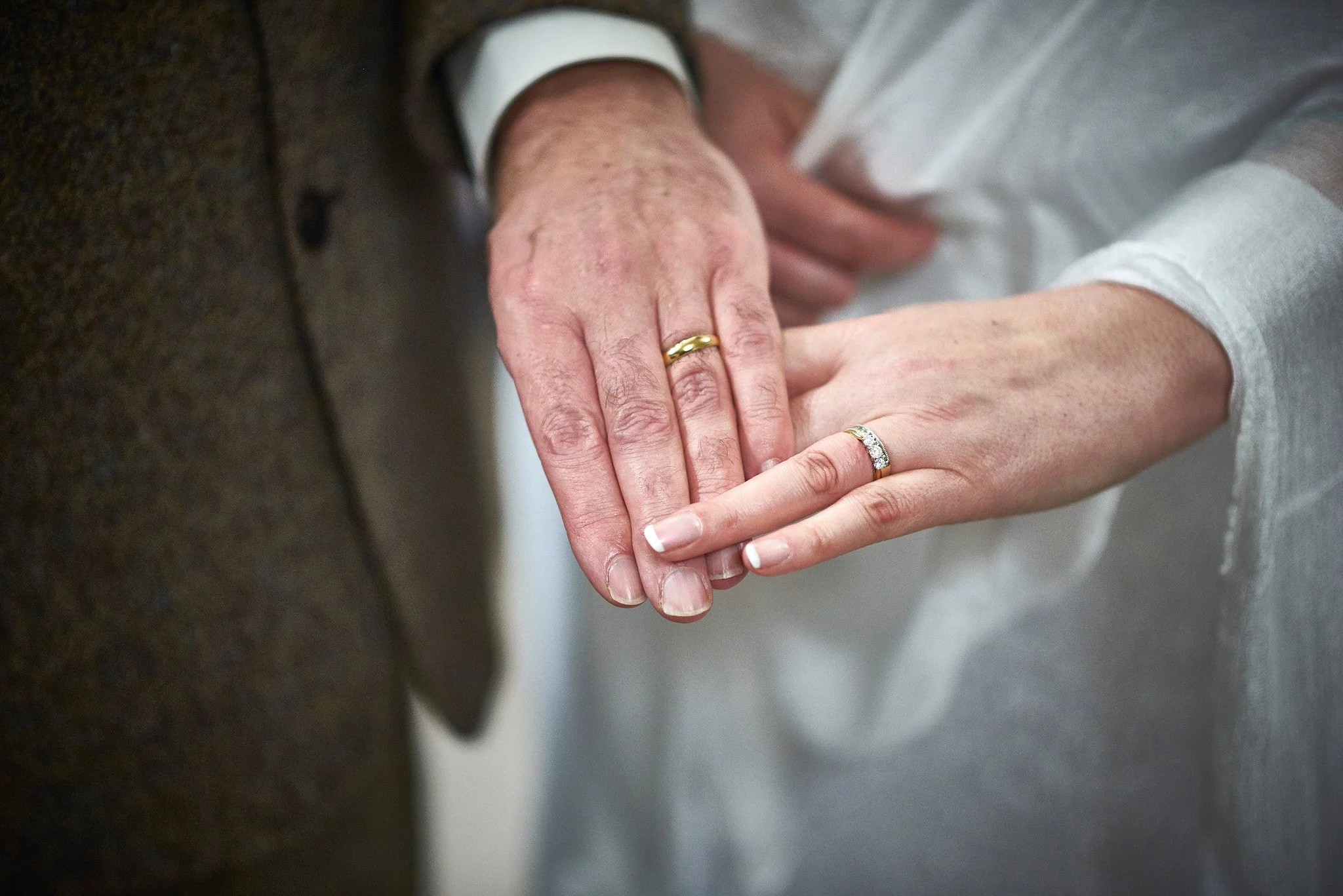Close-up of a couple's hands showing wedding rings, with the woman's hand on top of the man's. The woman has a silver ring with diamonds, and the man has a plain gold band. The background is blurred, focusing on the hands.