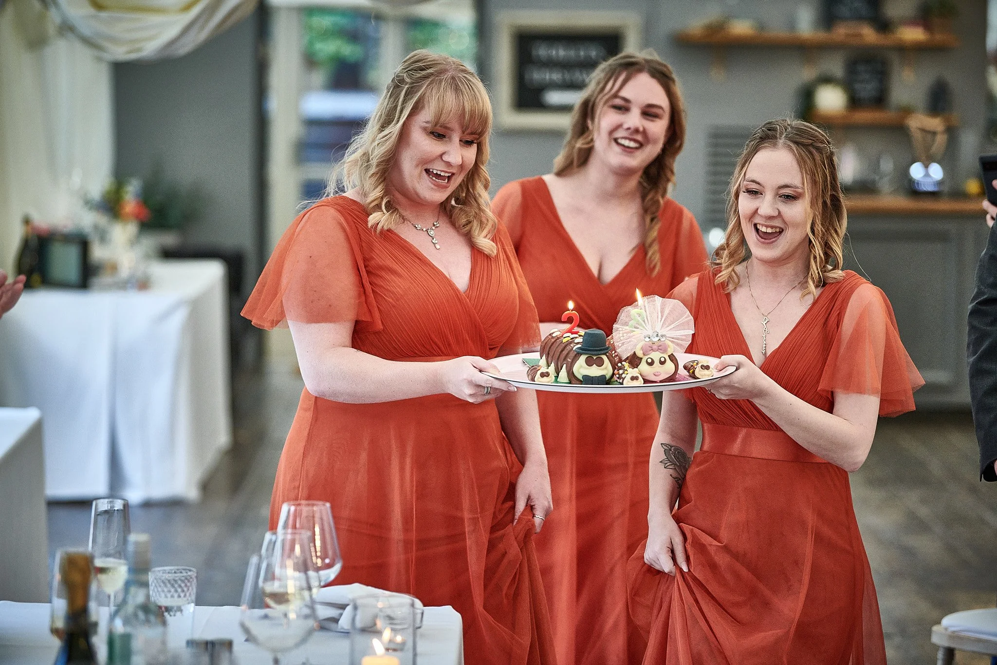 Three women in orange dresses holding a birthday cake with lit candles and decorated figures in a restaurant or banquet hall.