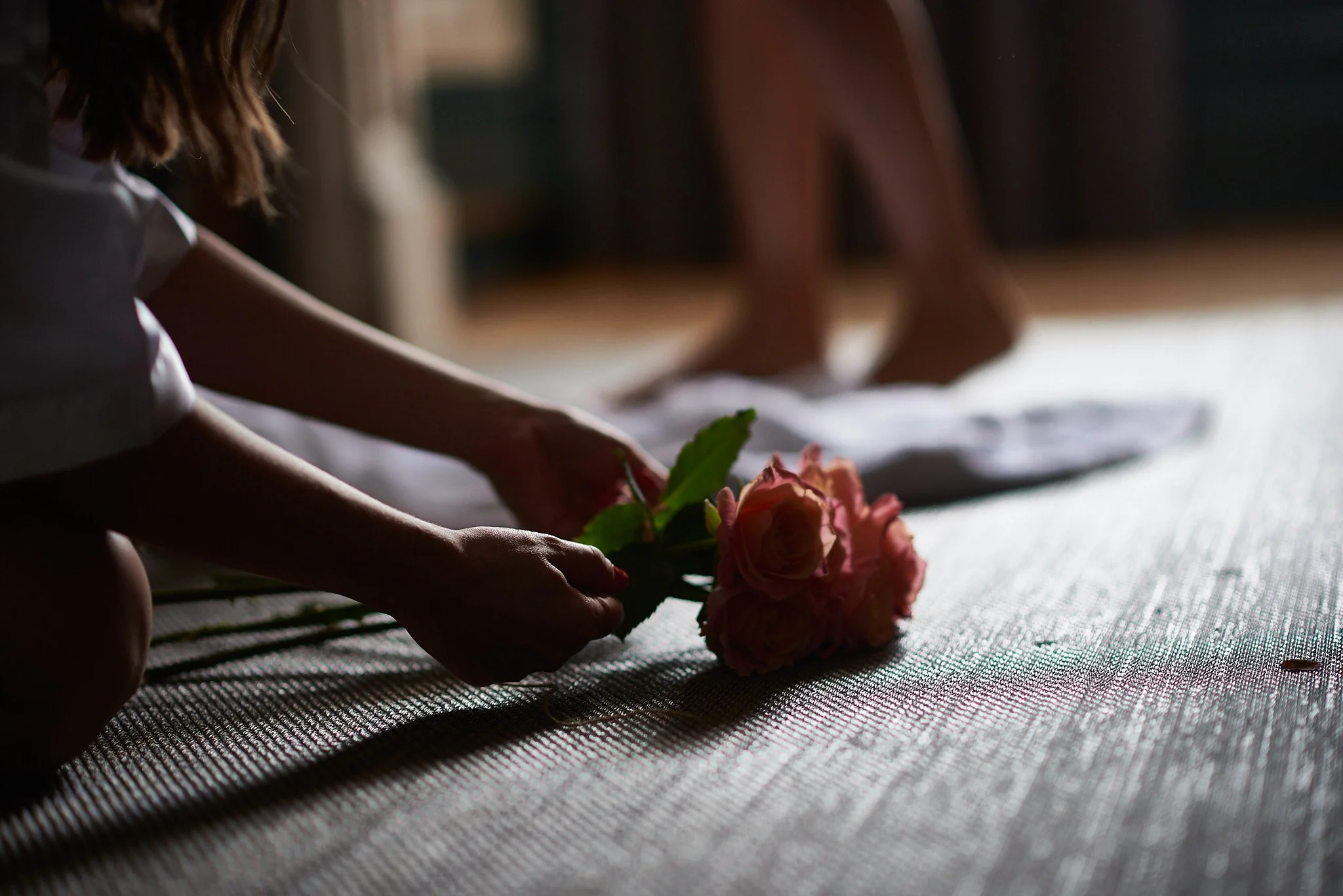 A young girl in white, kneeling on a textured surface, holding a bouquet of pink roses as sunlight filters through a window. In the background, a person stands on a white cloth.