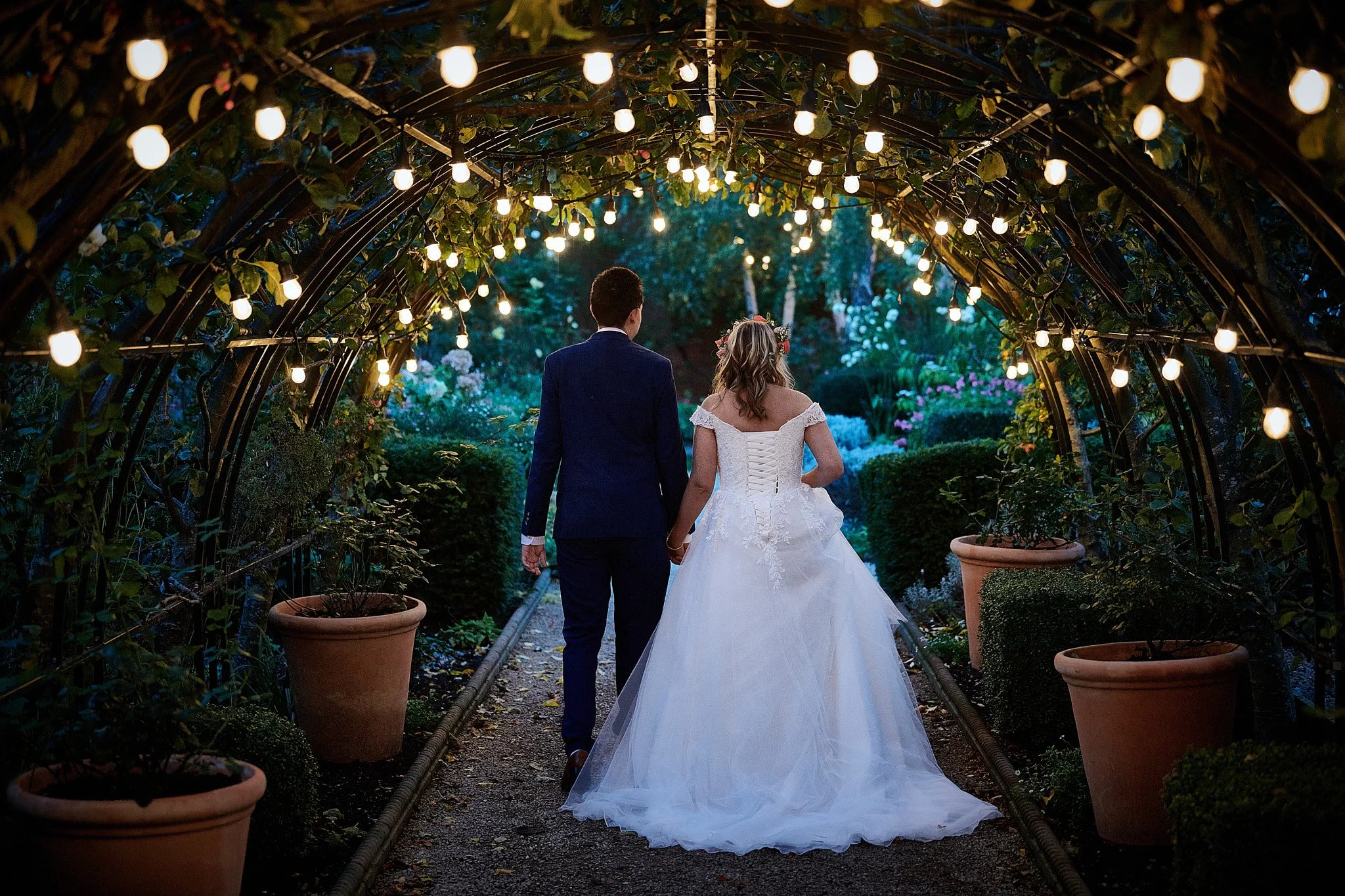 A newlywed couple walking hand-in-hand through a garden archway with string lights at dusk, surrounded by potted plants and lush greenery.