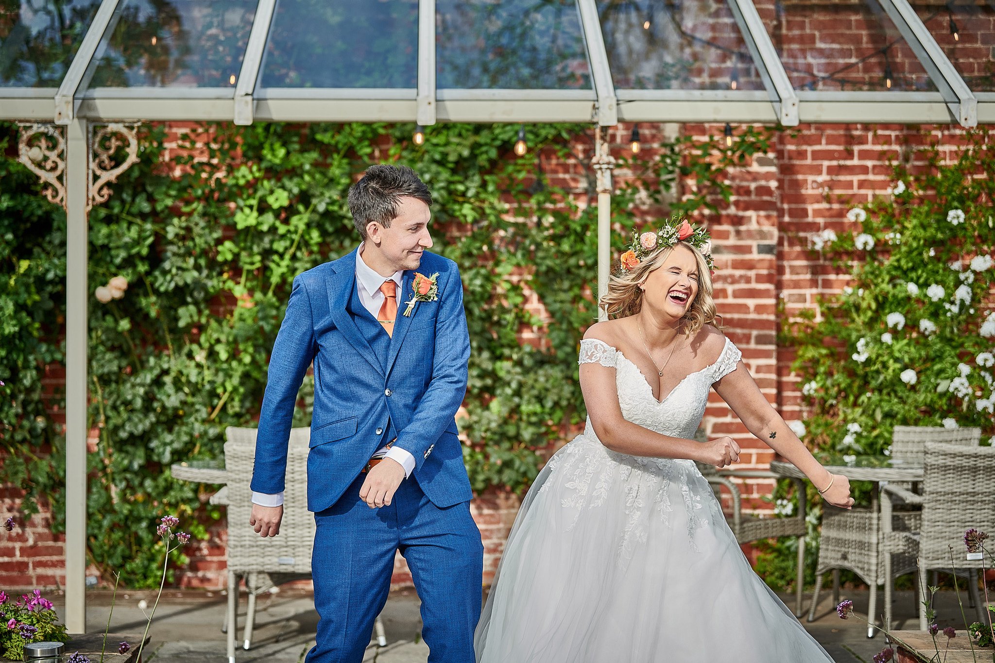 A newlywed couple dancing outdoors under a glass canopy, with the bride wearing a white off-the-shoulder wedding gown and a flower crown, and the groom in a blue suit, against a red brick wall and green foliage.