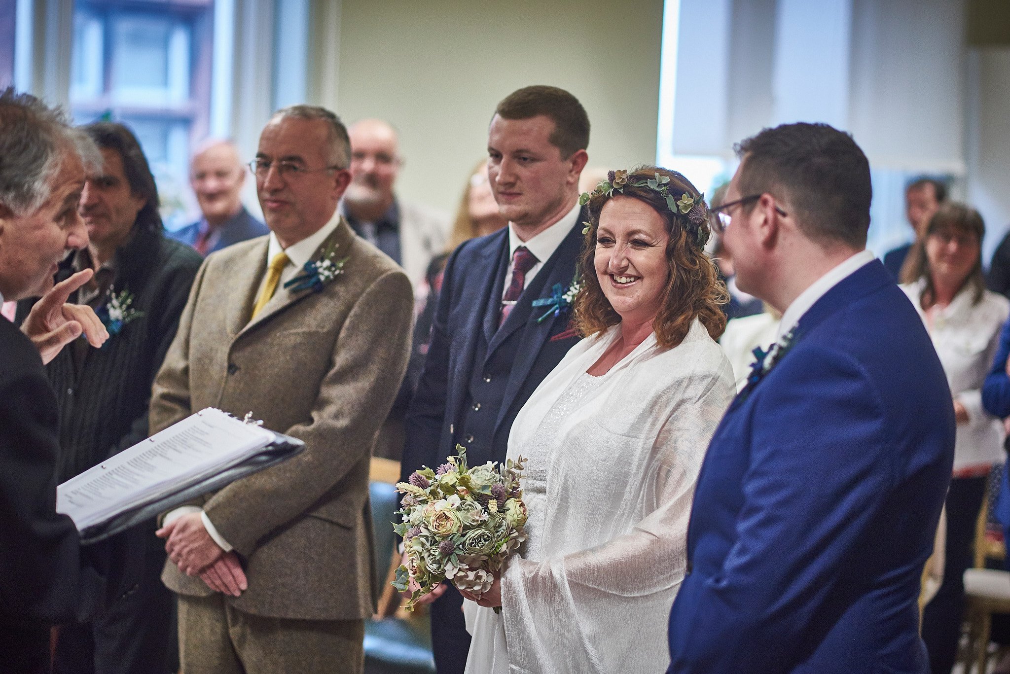 A bride holding a bouquet standing next to a groom during a wedding ceremony, surrounded by friends and family in an indoor setting.