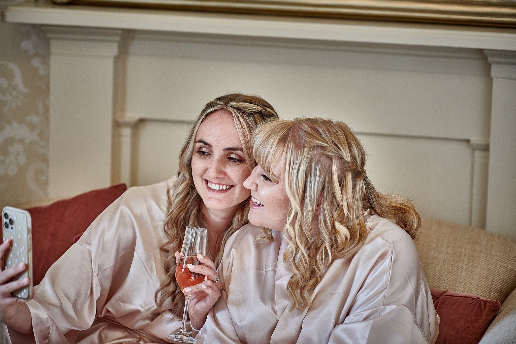 Two women in satin robes sitting on a couch, taking a selfie and smiling, one holding a glass of rosé wine.