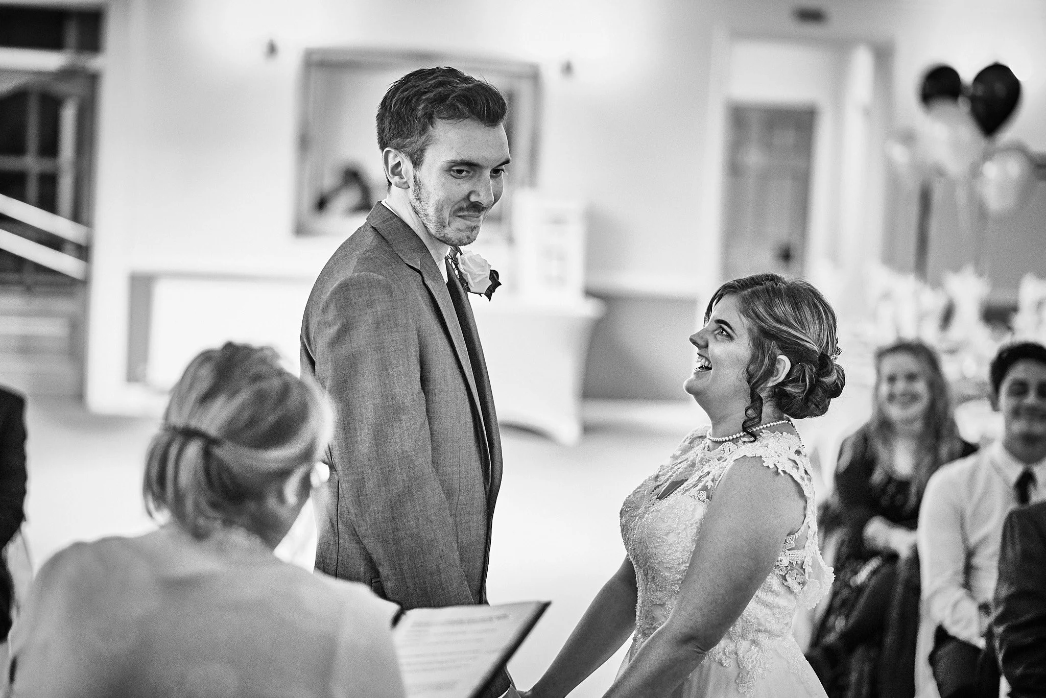 Black and white photo of a wedding ceremony with a bride and groom holding hands and looking at each other, surrounded by guests indoors.