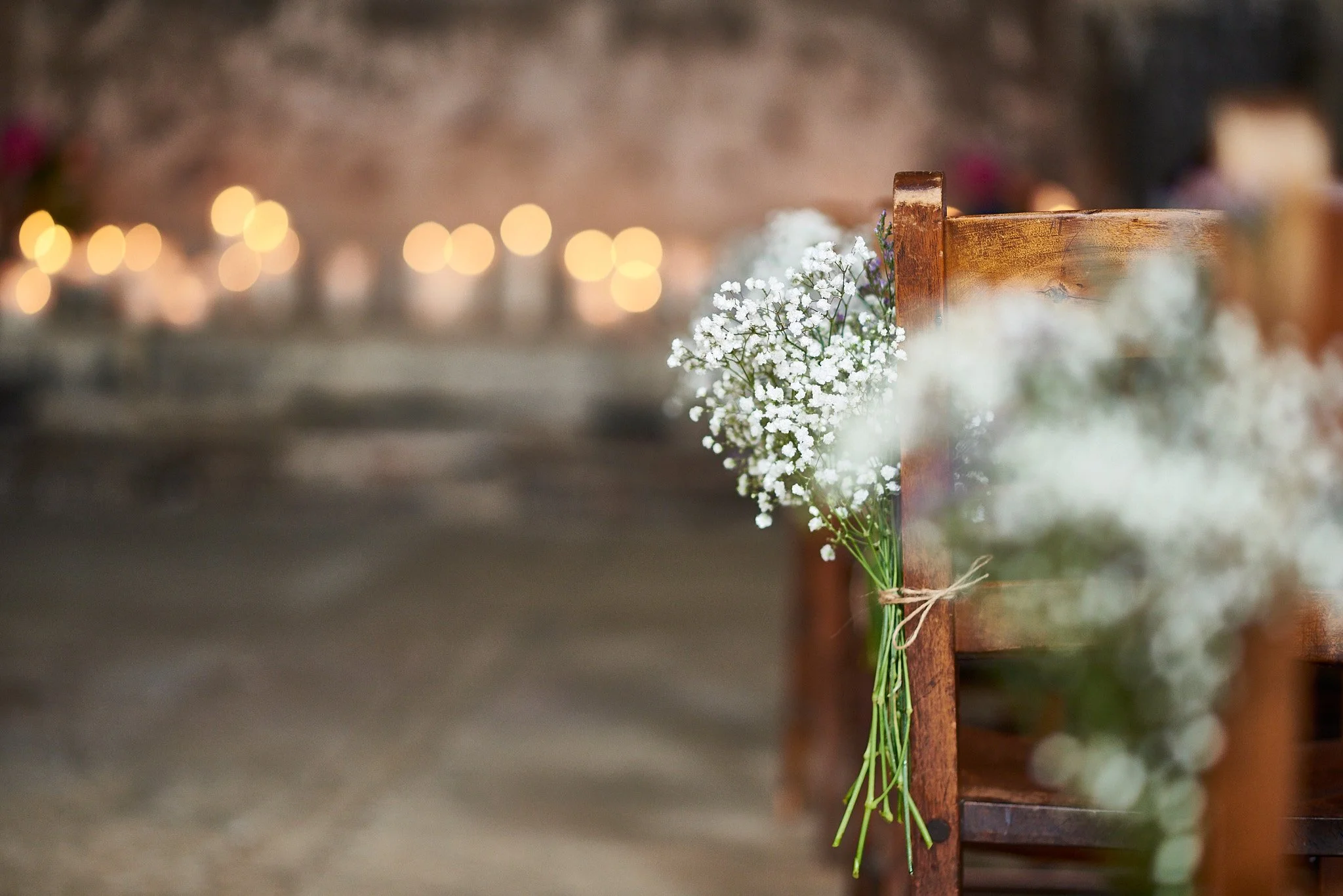 A small bouquet of white baby's breath flowers tied with twine is attached to the side of a wooden chair at a rustic indoor setting, with blurred warm lights in the background.