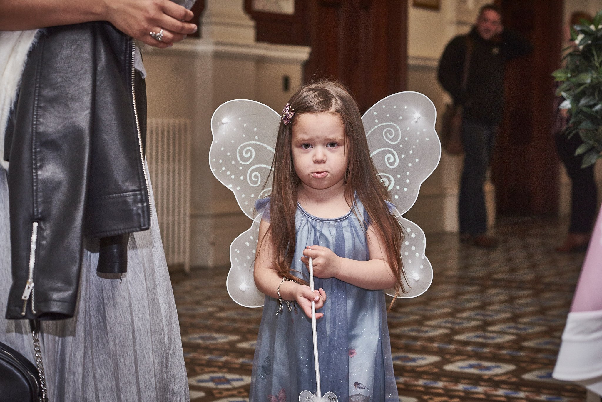 A young girl dressed as a fairy with translucent wings and a blue dress standing indoors, making a pouty face.