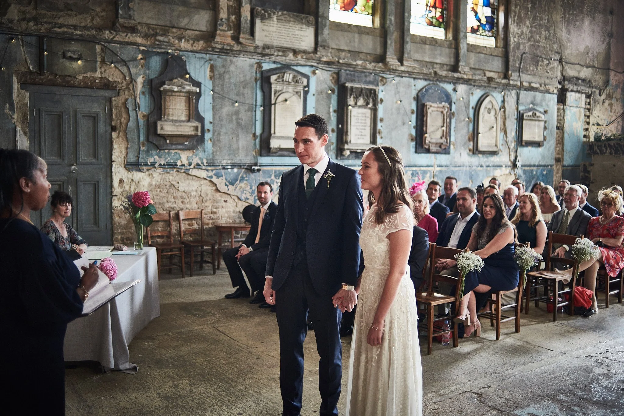 A wedding ceremony taking place in an old, rustic church with weathered walls and stained glass windows. The bride and groom stand hand in hand facing the officiant, with guests seated behind them.