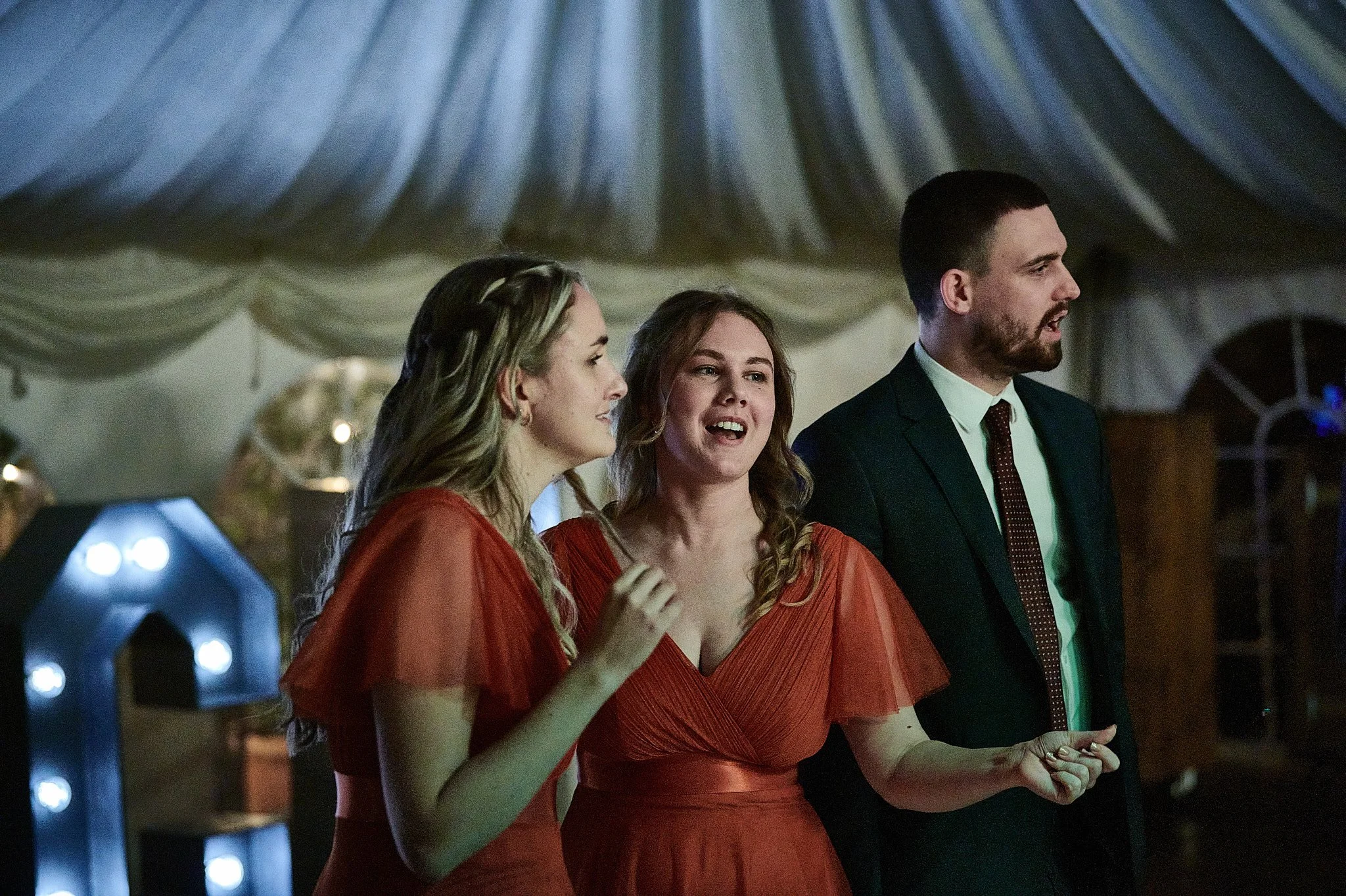 Three people, two women in red dresses and a man in a suit, are standing together and talking at an indoor event.