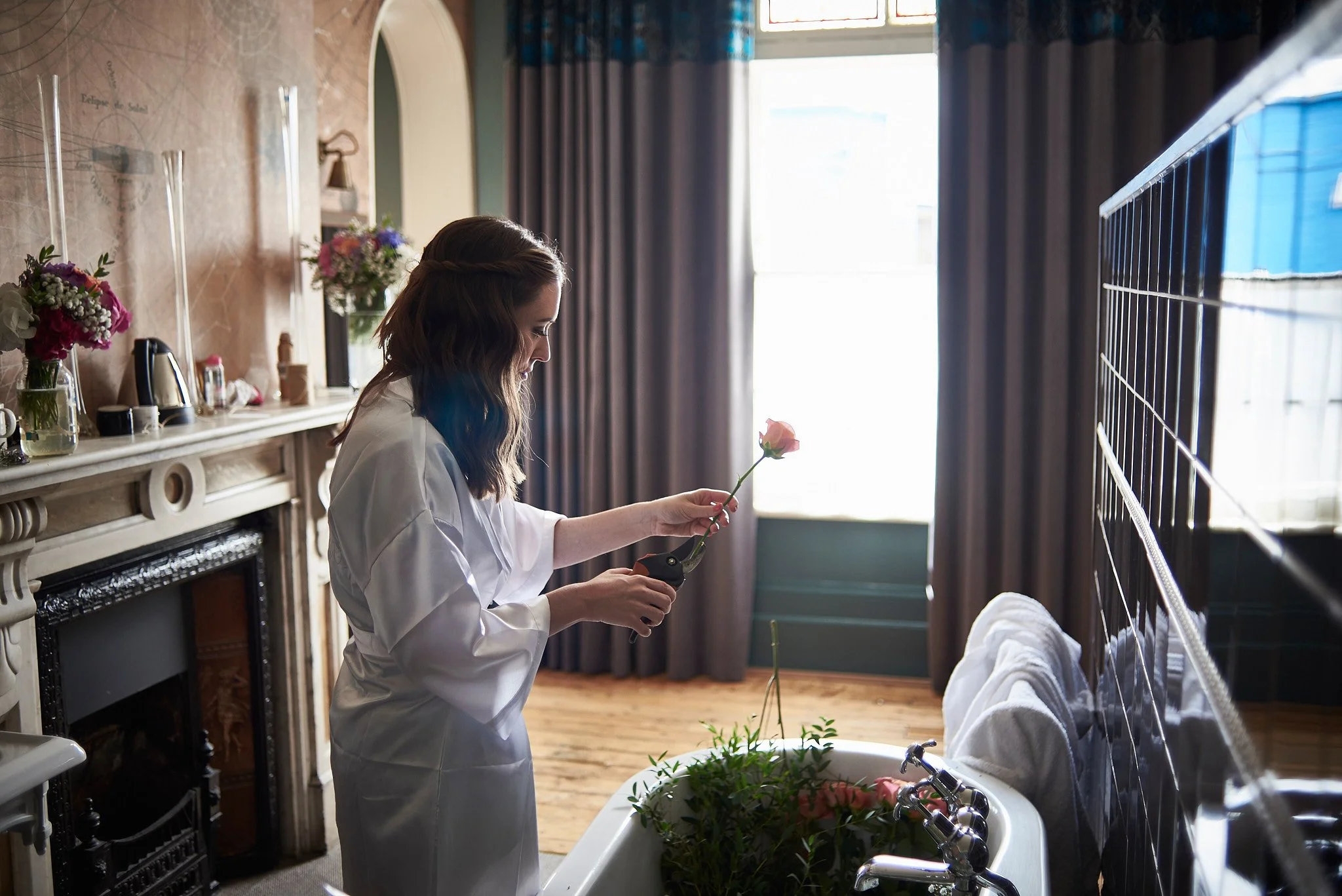 A woman in a white robe watering a pink flower in a bathtub filled with plants.