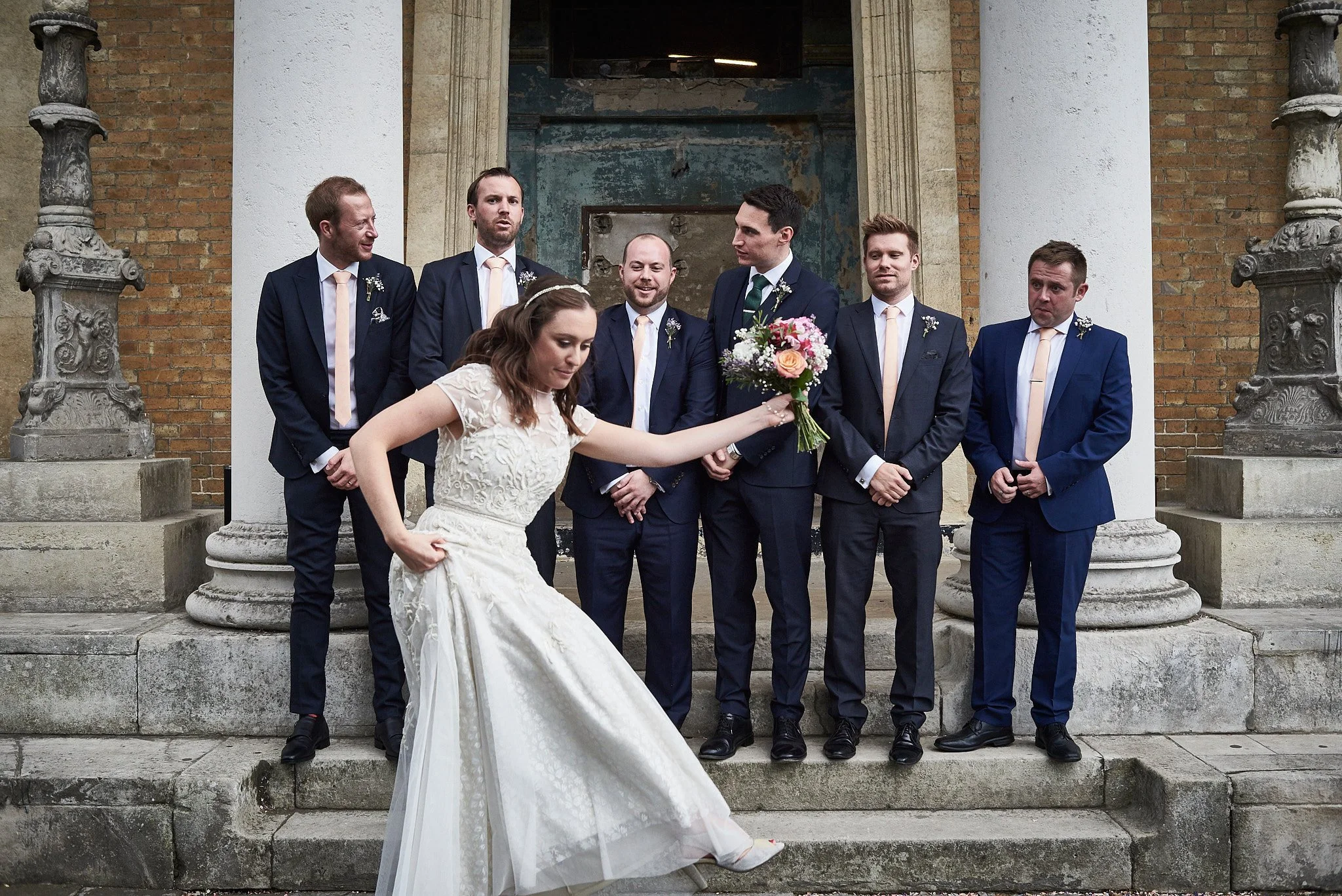 A woman in a white wedding dress holding a bouquet of flowers, standing in front of a group of men dressed in suits outside a building with columns.