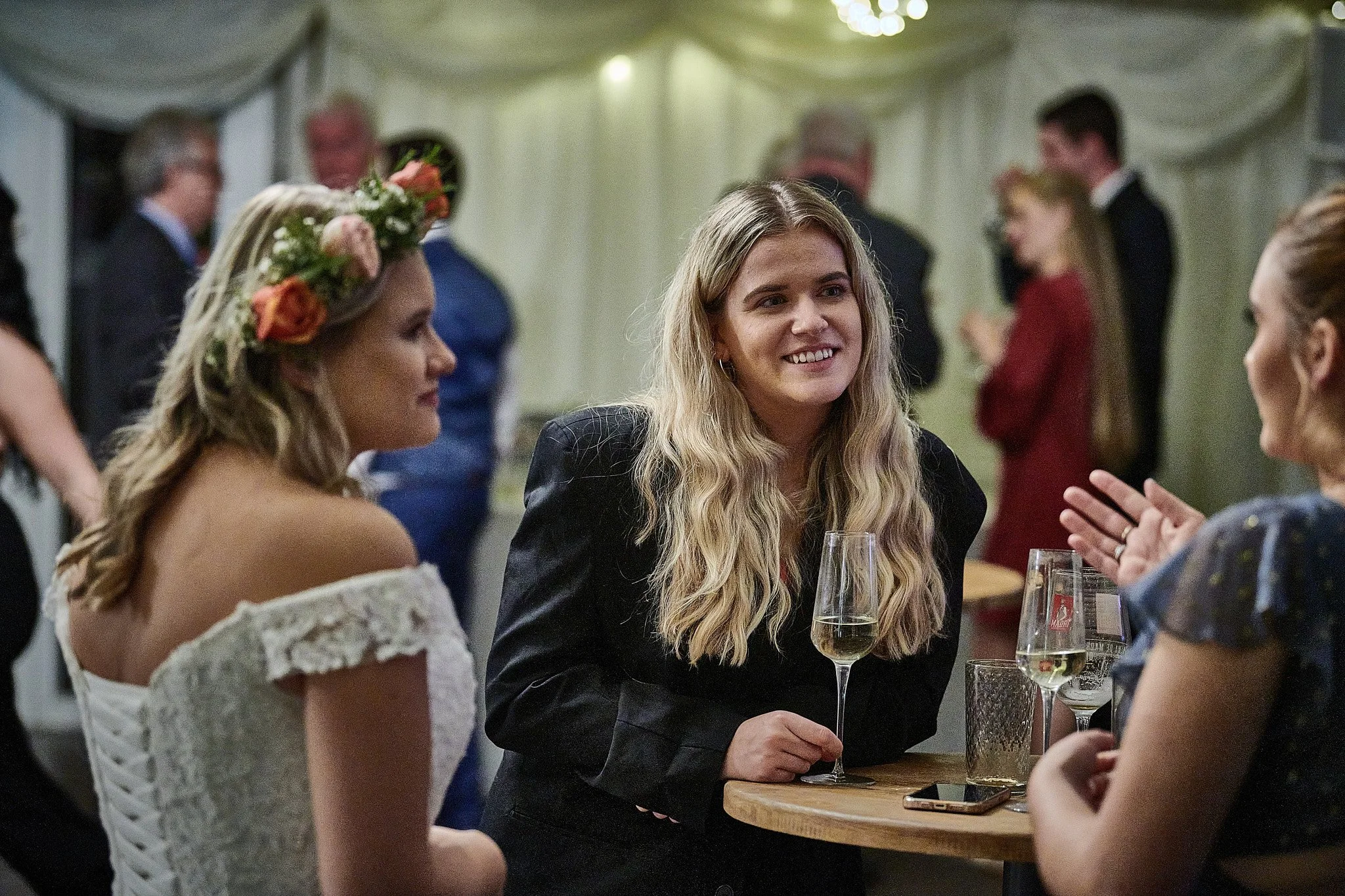 Three women are seated at a round table at a social gathering. The woman on the left wears a white off-the-shoulder dress and a floral crown, and the woman in the middle wears a black blazer, smiling and holding a glass of champagne. The woman on the