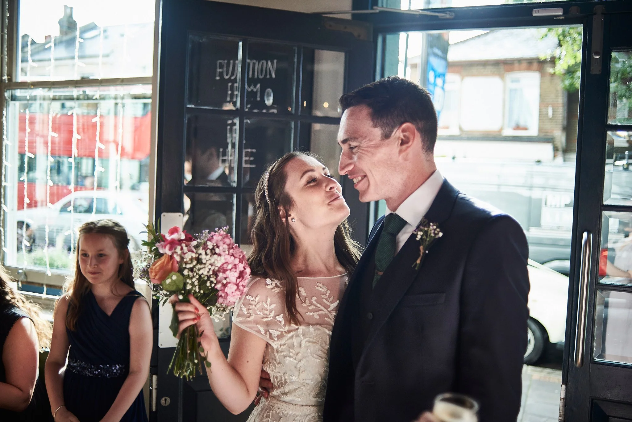 A bride and groom share a joyful moment at their wedding reception. The bride holds a bouquet of pink and white flowers and wears a white lace dress. The groom is in a black suit with a white shirt and green tie. In the background, a young girl in a 