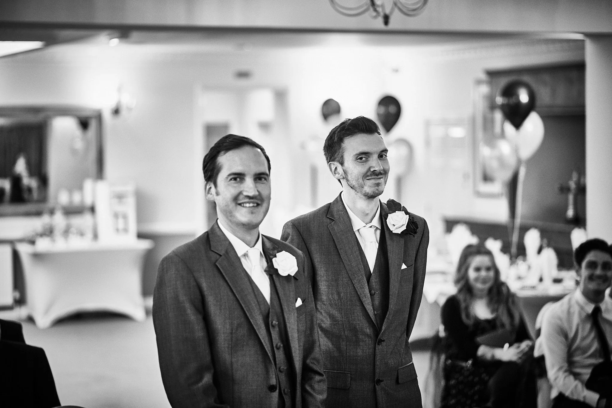 Two men in suits with boutonnières, standing side by side at a wedding reception, smiling while guests watch in the background.