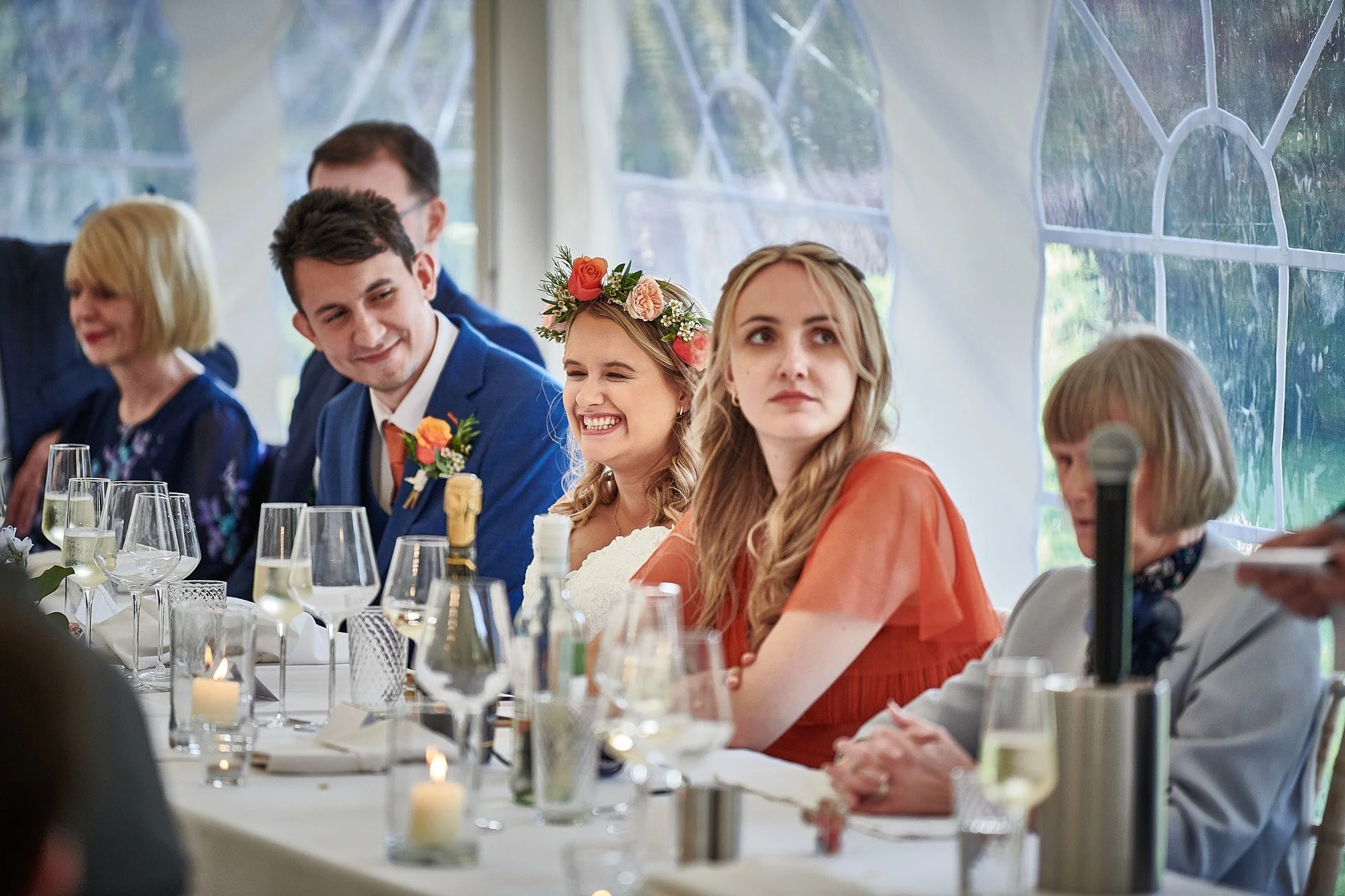 Group of people at a wedding reception, sitting at a table with glasses and candles, inside a tent with large windows.