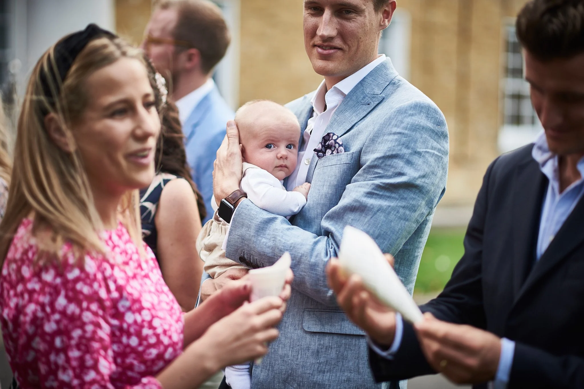 A man in a light blue suit holding a baby at an outdoor event surrounded by smiling adults.