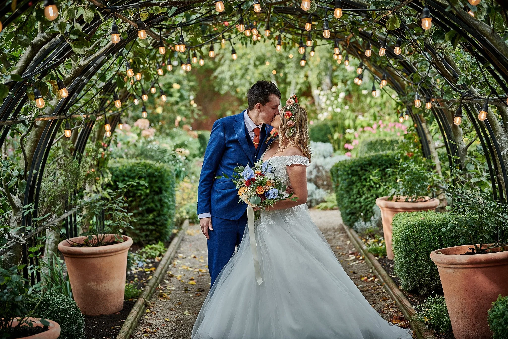 Wedding couple kissing under a floral archway with hanging lights in a lush garden.