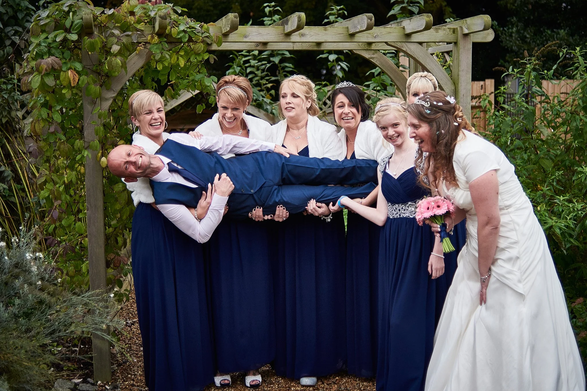 A group of women in formal dresses lifting a man in a suit in a garden setting, smiling and laughing.