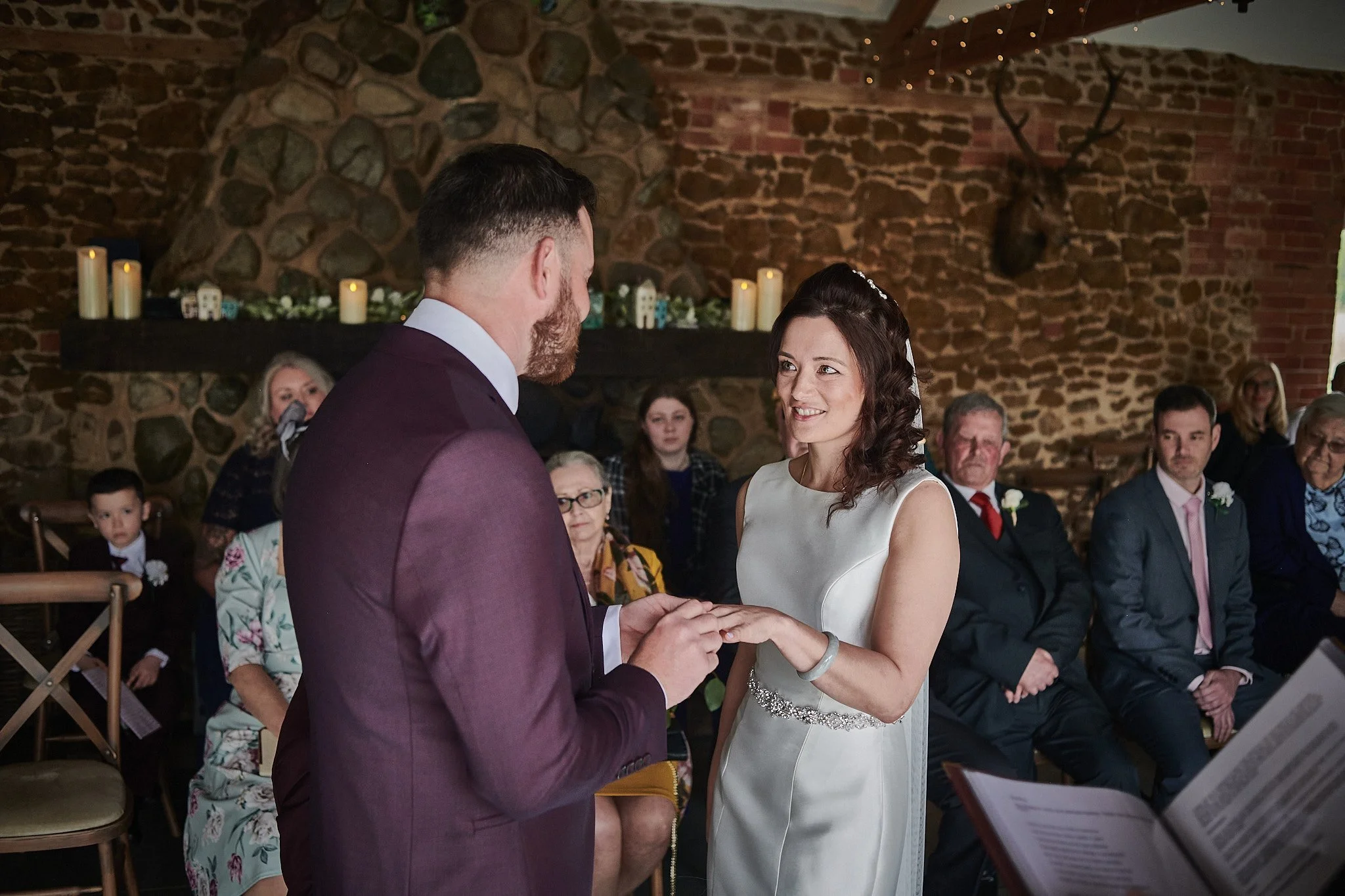 A couple exchanging wedding rings during a wedding ceremony in a rustic venue with a brick and stone wall, surrounded by seated guests.