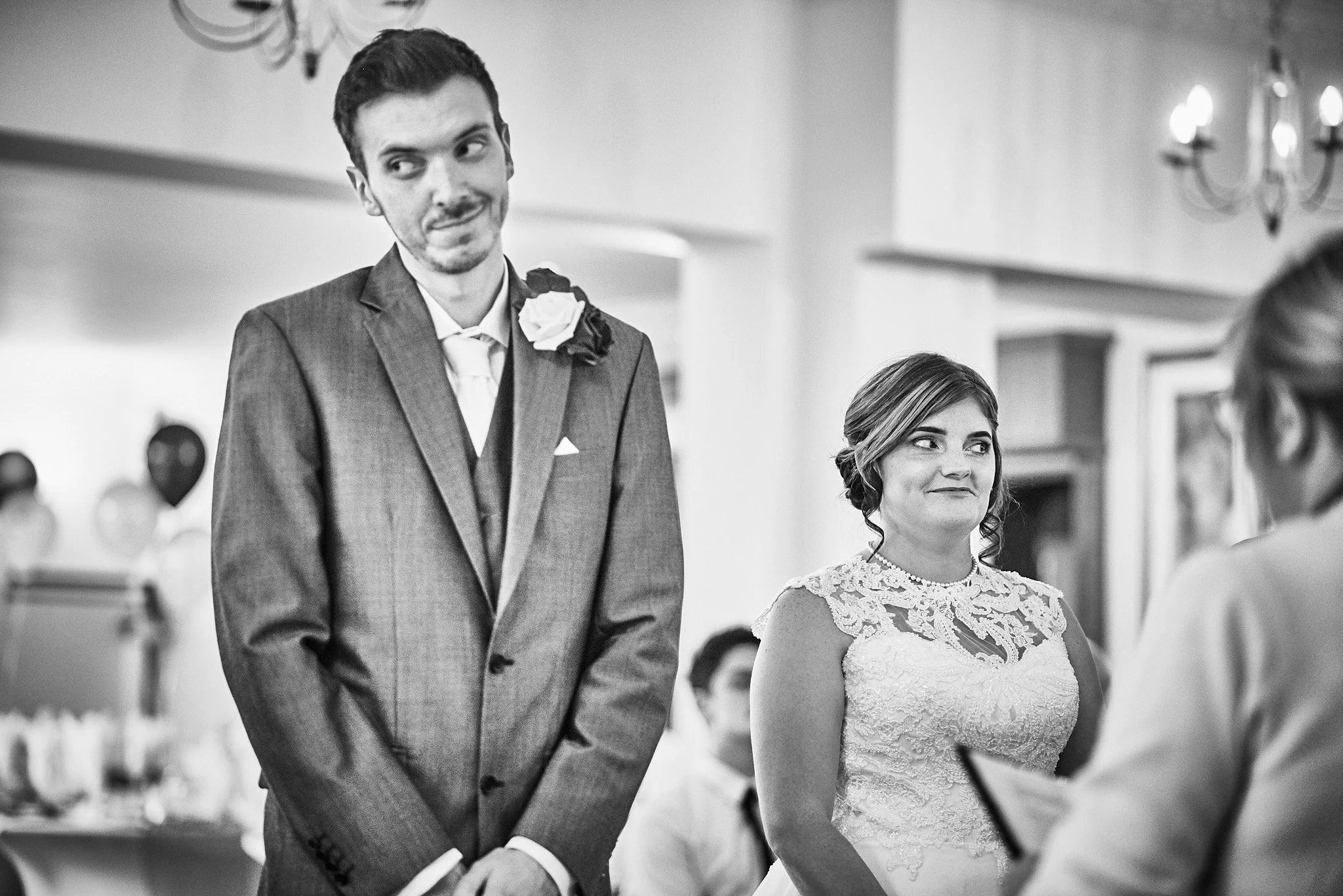 A black and white photo of a wedding ceremony showing a bride and groom standing and listening to an officiant. The groom is wearing a suit with a boutonniere, and the bride is wearing a lace wedding dress with a necklace.