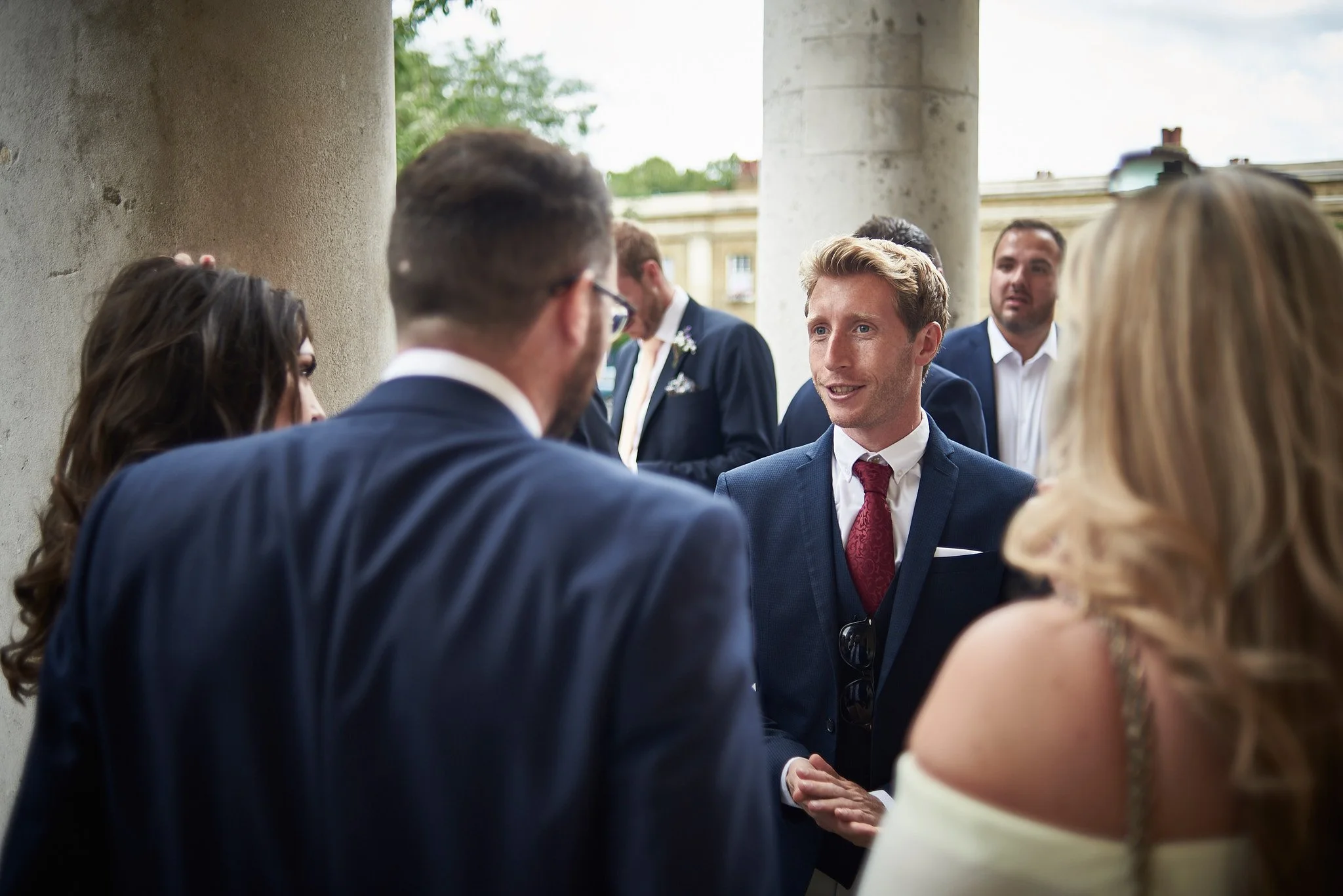 Group of people in formal attire socializing outdoors, with one man in a blue suit and red tie speaking to others.