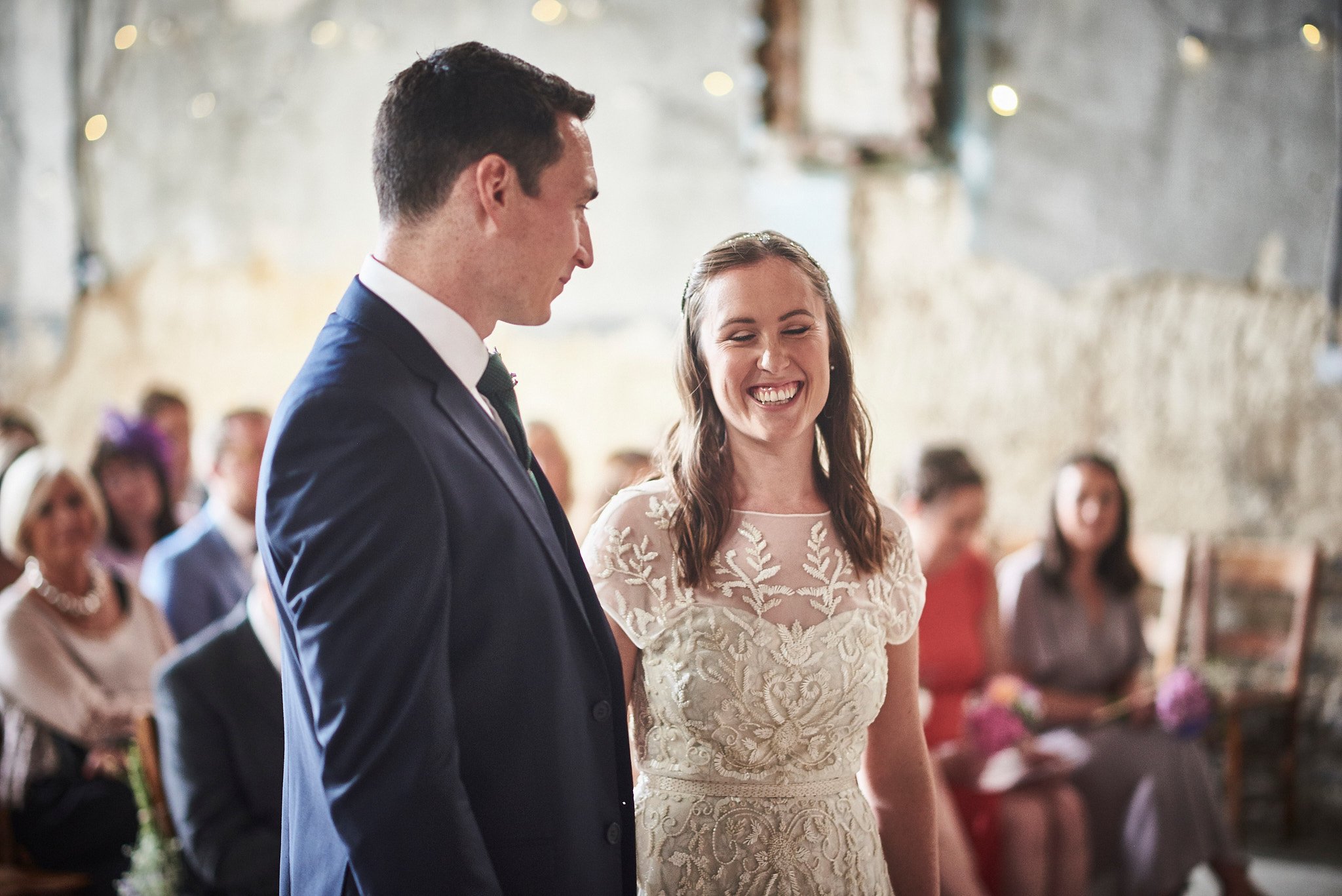A bride and groom standing at their wedding ceremony in a rustic venue, smiling, with guests seated behind them.
