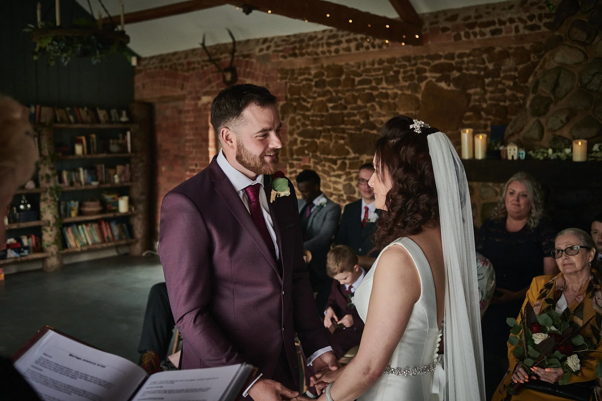 Bride and groom holding hands and gazing at each other during their wedding ceremony indoors with family and friends watching.