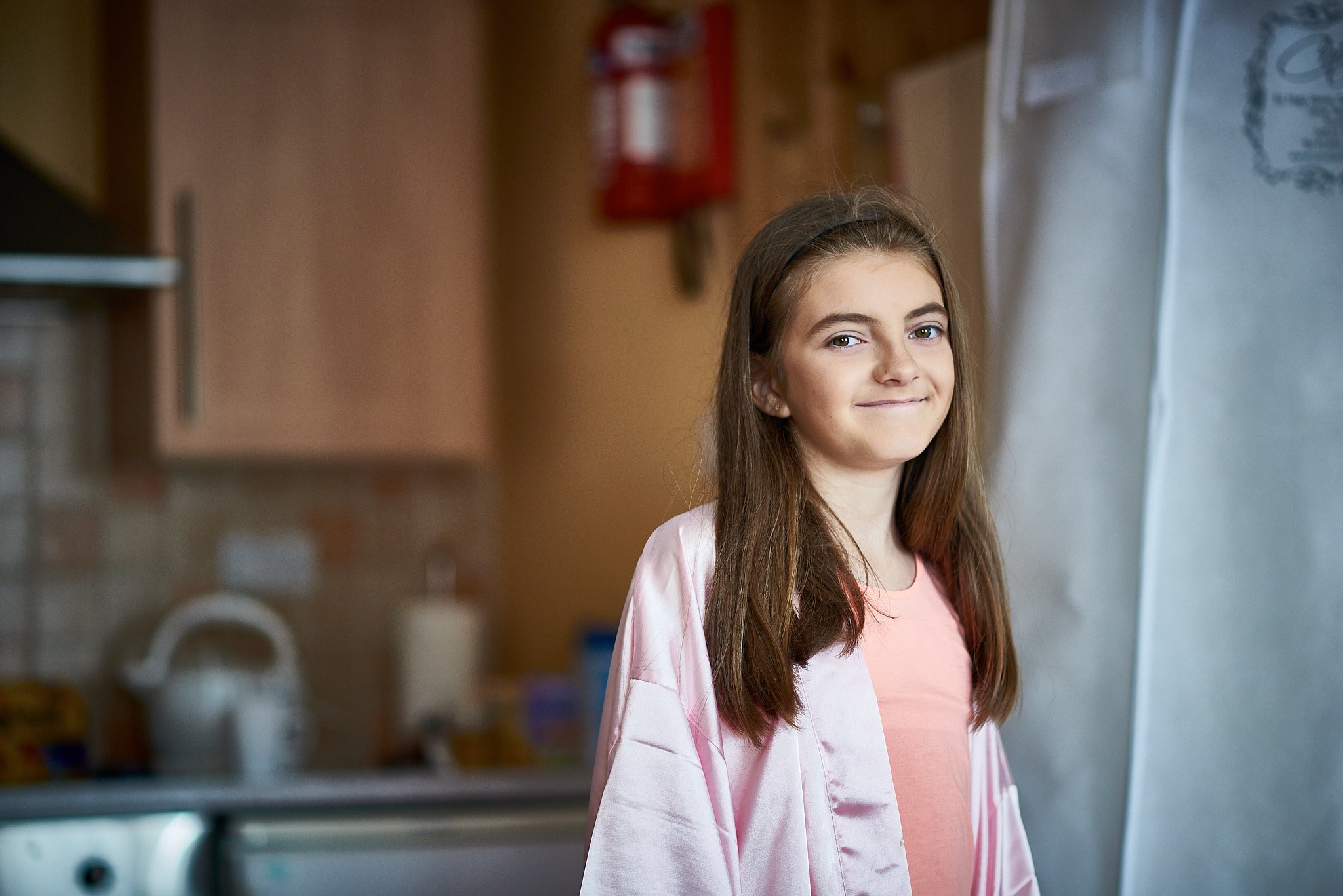 A young girl with long brown hair, wearing a pink robe over a peach shirt, standing in a kitchen and smiling slightly.