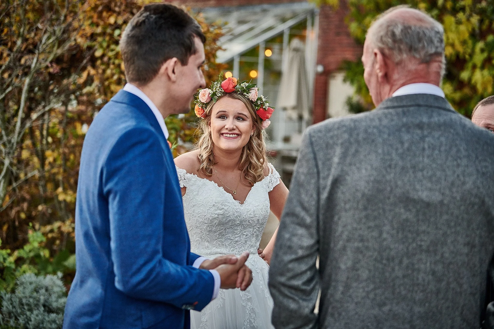 Bride and groom smiling during outdoor wedding ceremony, with officiant present, autumn foliage background.