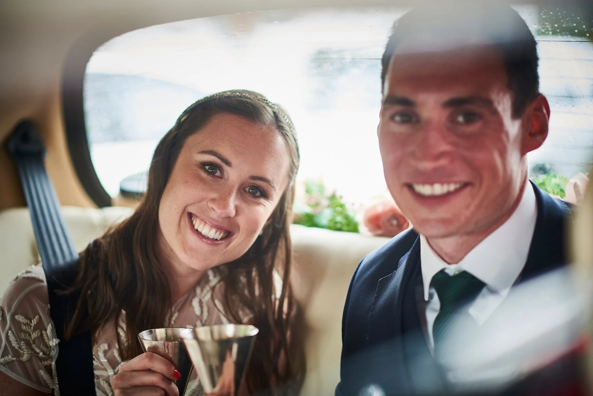 A smiling woman and man are seen through the rear window of a vehicle, holding glasses of drinks and dressed formally, likely at a celebration or wedding.