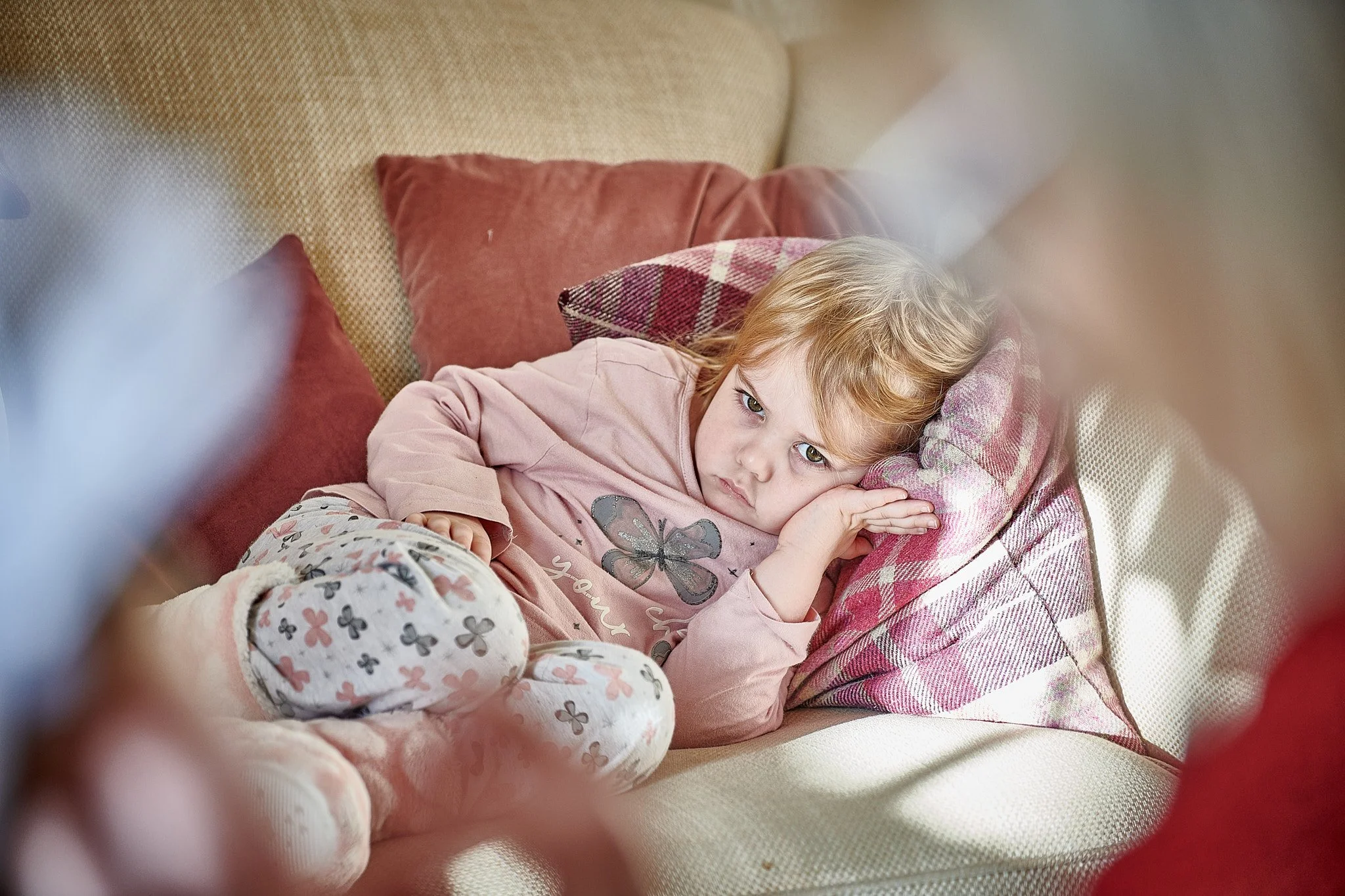 A young girl with blond hair lying on a beige couch with pink and plaid pillows, resting her head on her hand and looking at the camera.