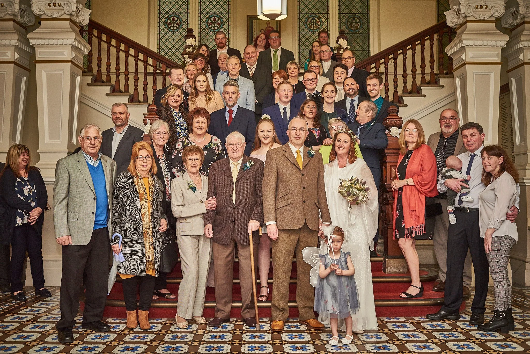 Group of people gathered on a staircase in a historic building, celebrating a wedding, with most dressed in formal attire, and a woman holding a bouquet of flowers.
