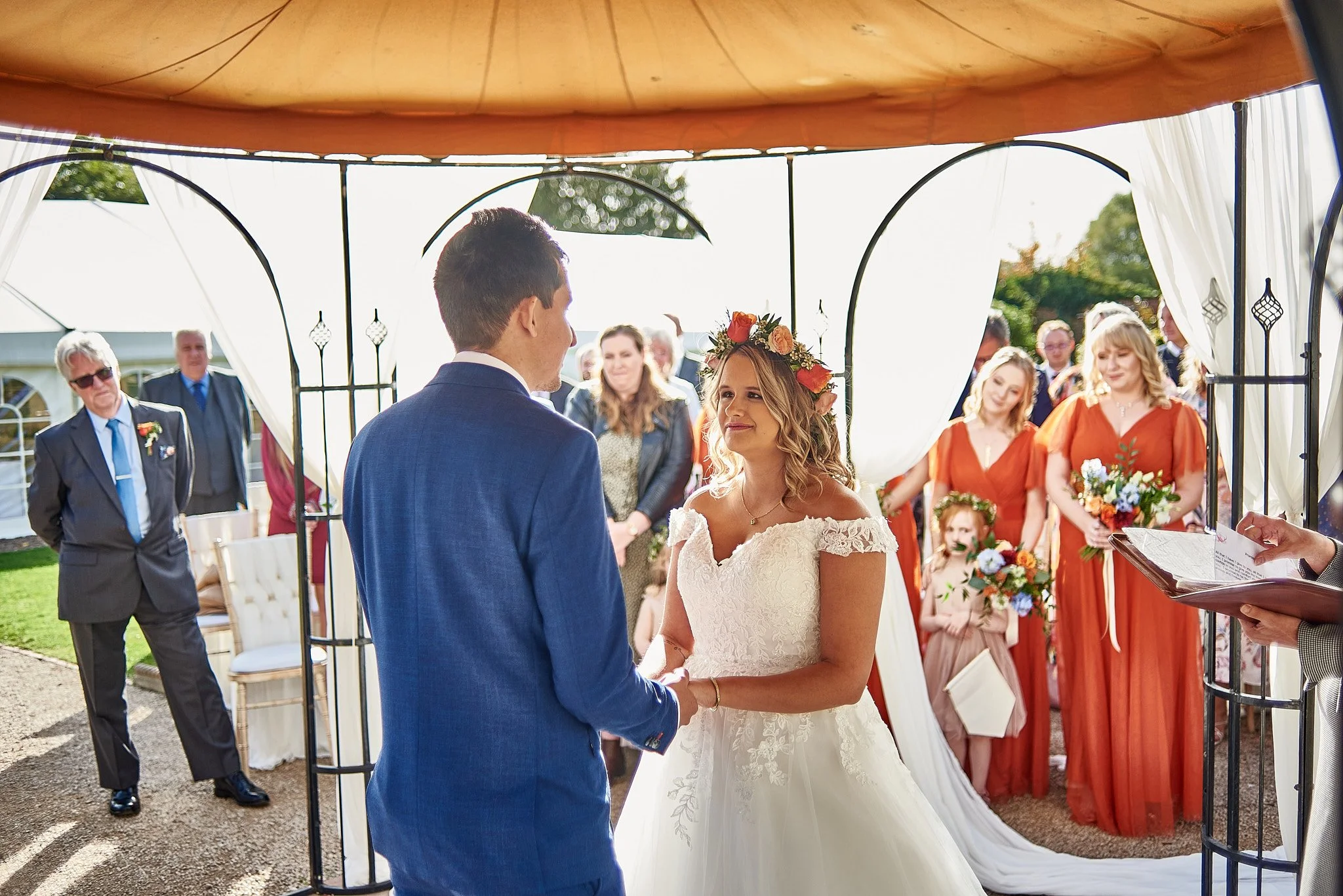 A bride and groom exchanging vows at an outdoor wedding ceremony under a decorated canopy, with bridesmaids in orange dresses and guests watching.
