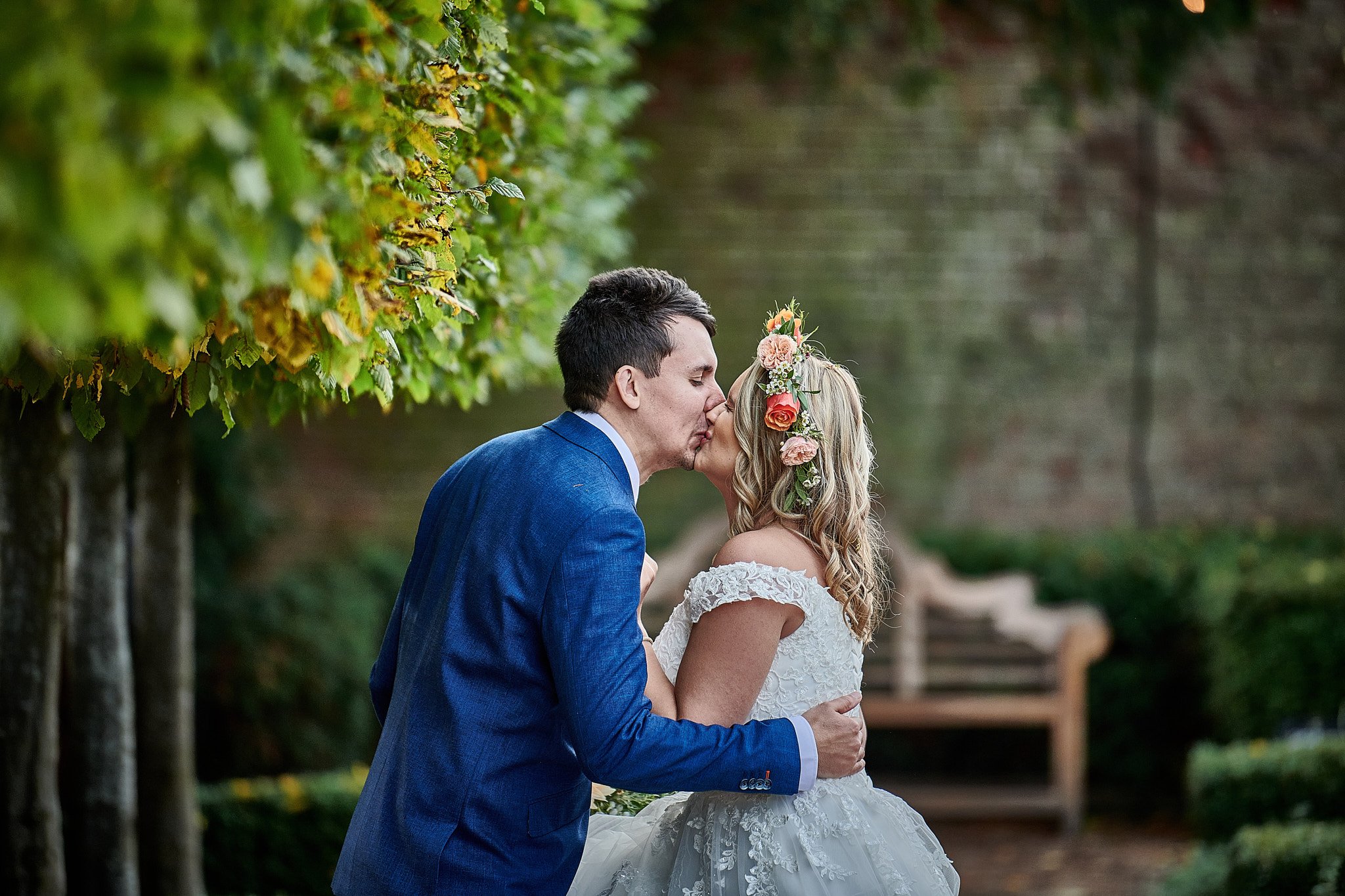 A bride and groom kiss outdoors on their wedding day, with the bride wearing a floral crown and the groom in a blue suit, surrounded by green foliage and a park bench in the background.