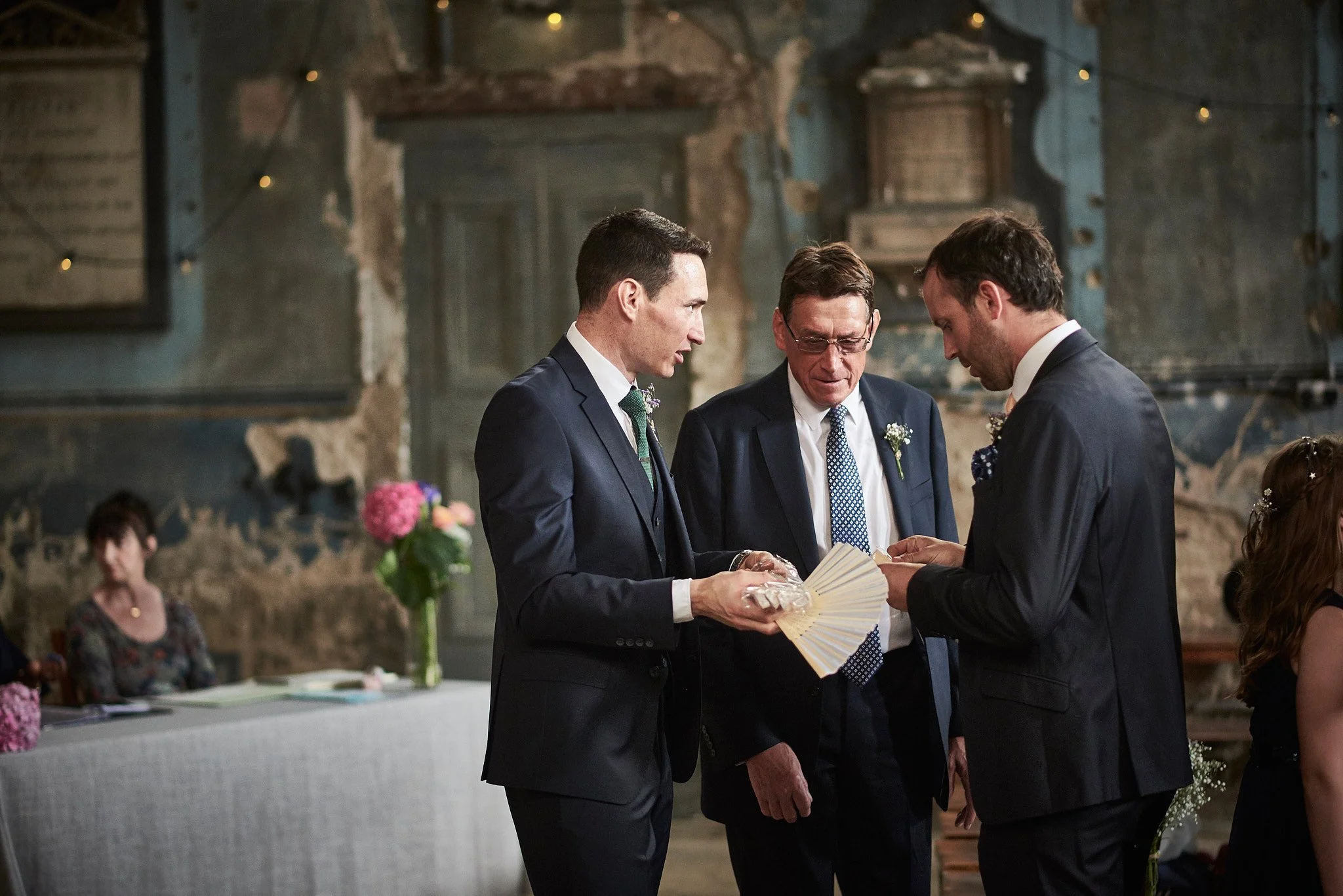 Two men in suits exchanging rings during a wedding ceremony, with a officiant standing between them holding a fan and a small box, in a rustic venue with onlookers seated at a table decorated with pink flowers.