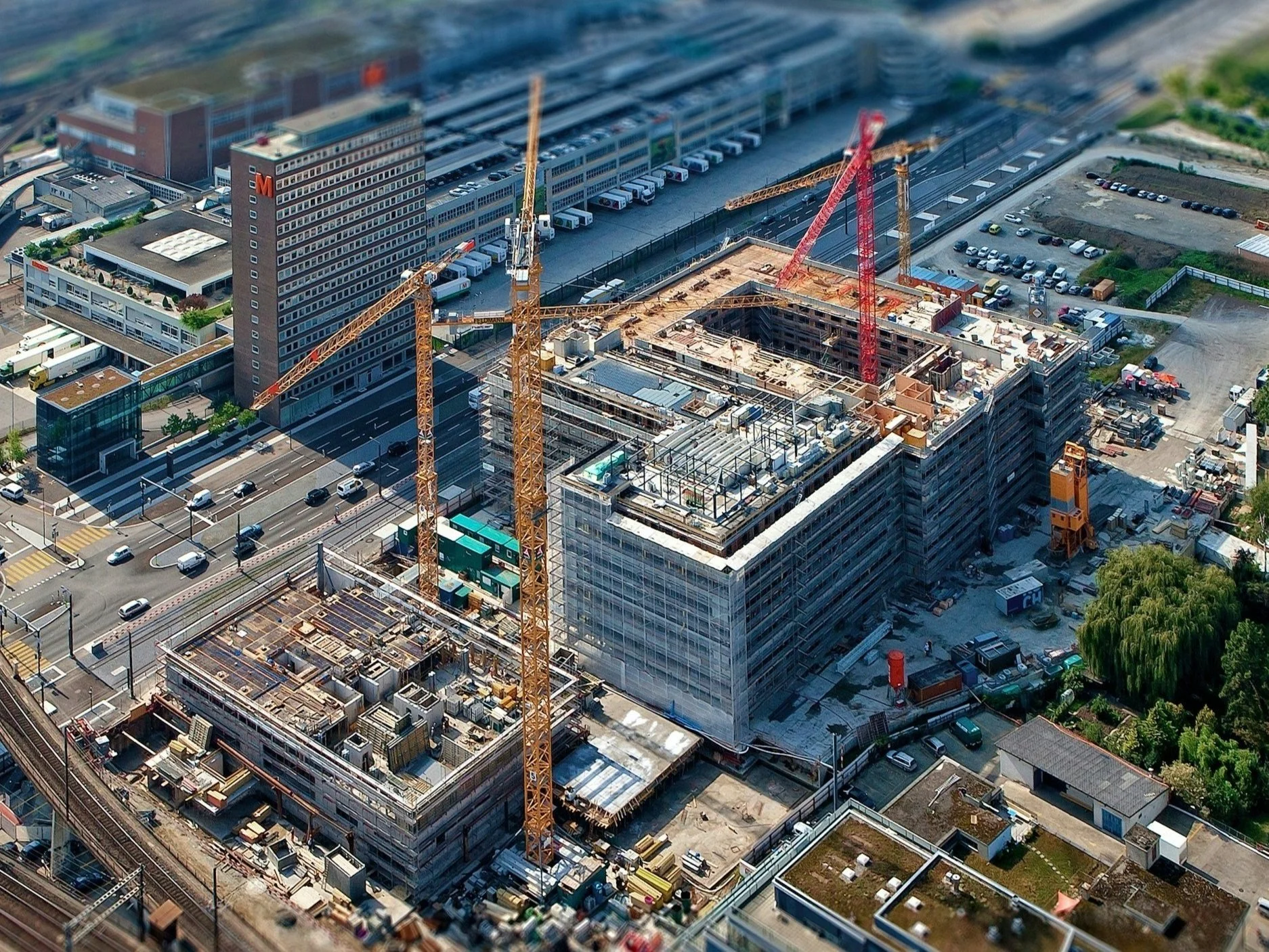 Bauarbeiten an einem großen Gebäude mit mehreren Baukränen, umgeben von Straßen, Parkplätzen und mehreren Gebäuden in einer Stadt.
