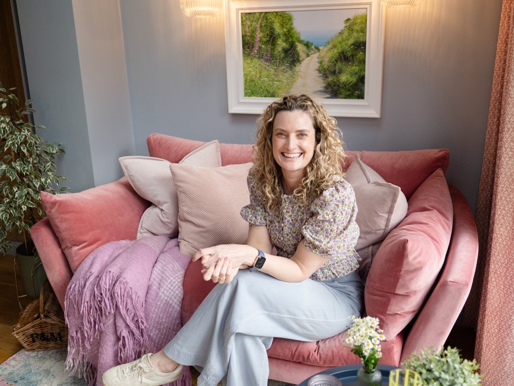 A woman with curly hair sitting on a pink sofa in a cozy living room, smiling at the camera. The sofa has light and dark pink pillows and a purple blanket. There is a painting of a scenic nature path hanging on the wall behind her. A potted plant is visible on the left. A small table with a plant and a yellow decorative item is in the foreground.