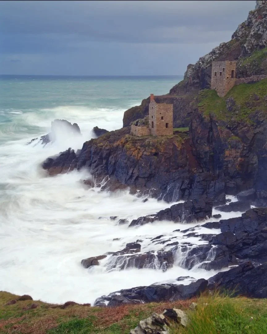 A rugged coastline with waves crashing against dark rocks, and old stone structures built on the cliffs.