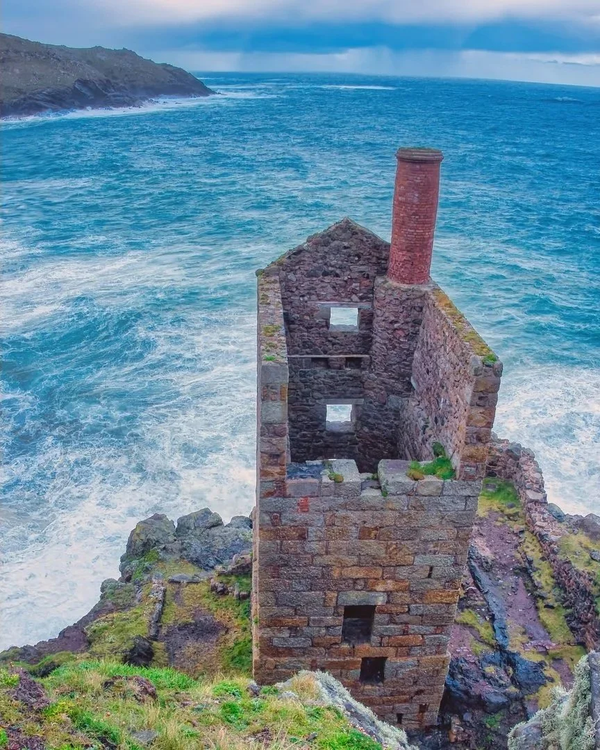A dilapidated stone building with a tall brick chimney, situated on a cliff overlooking the ocean with waves crashing against the rocks.