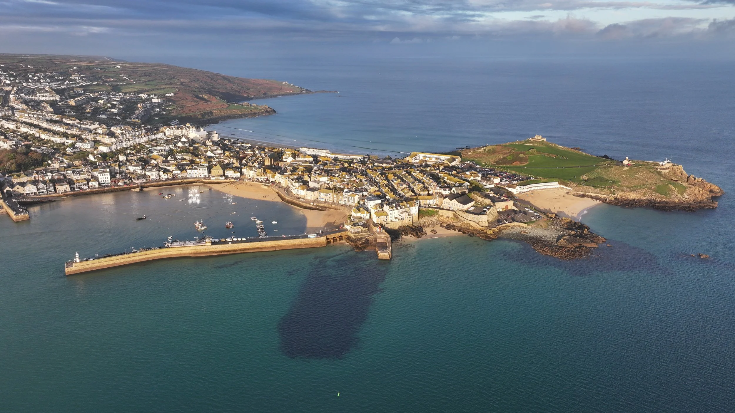 Aerial view of a coastal town with a harbor, sandy beaches, and houses lining the shore next to the ocean, with some green hills and a cloudy sky.