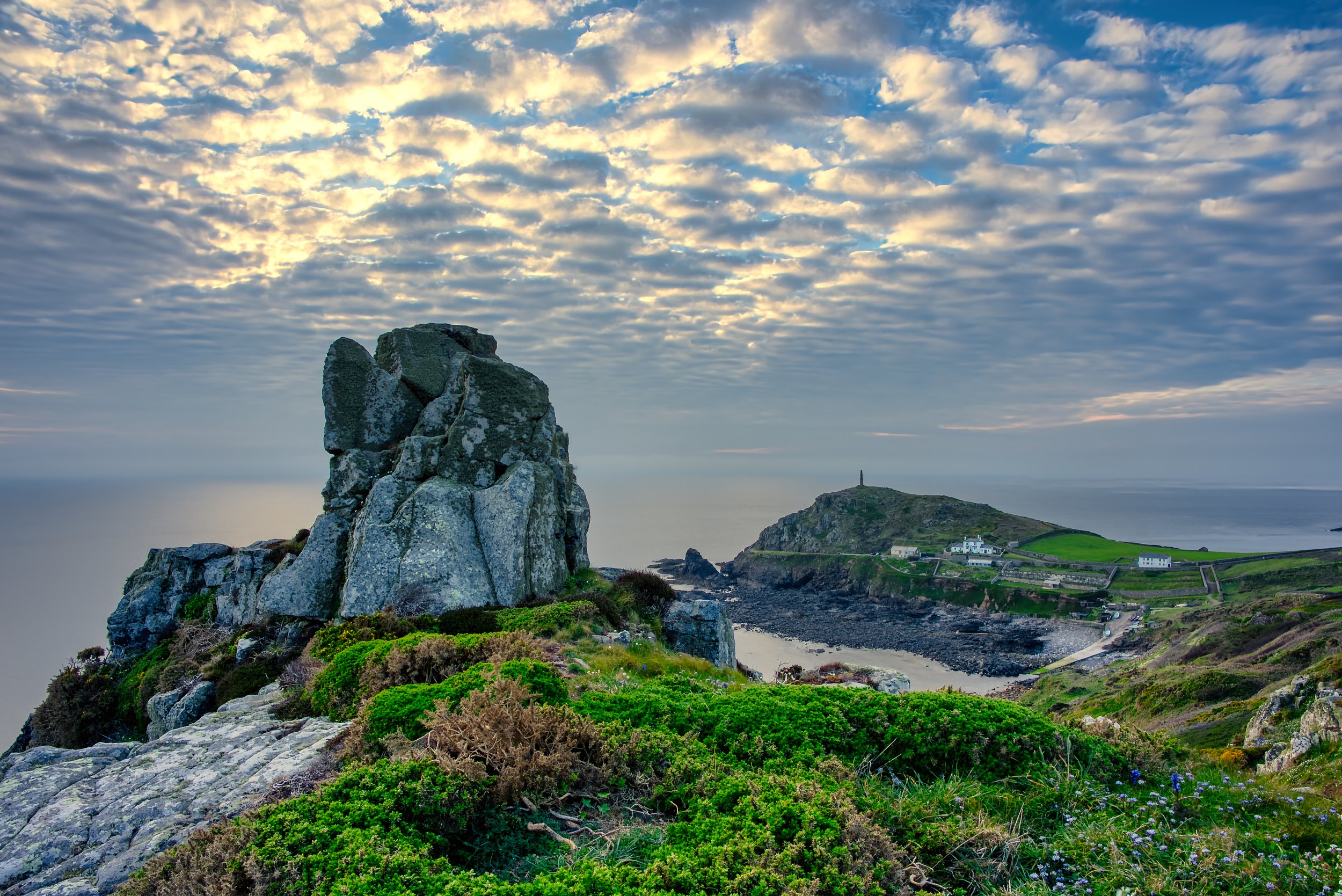 View over Cape Cornwall from the cliffs, showing granite rocks in the foreground, the coastal headland, sea and dramatic evening clouds.
