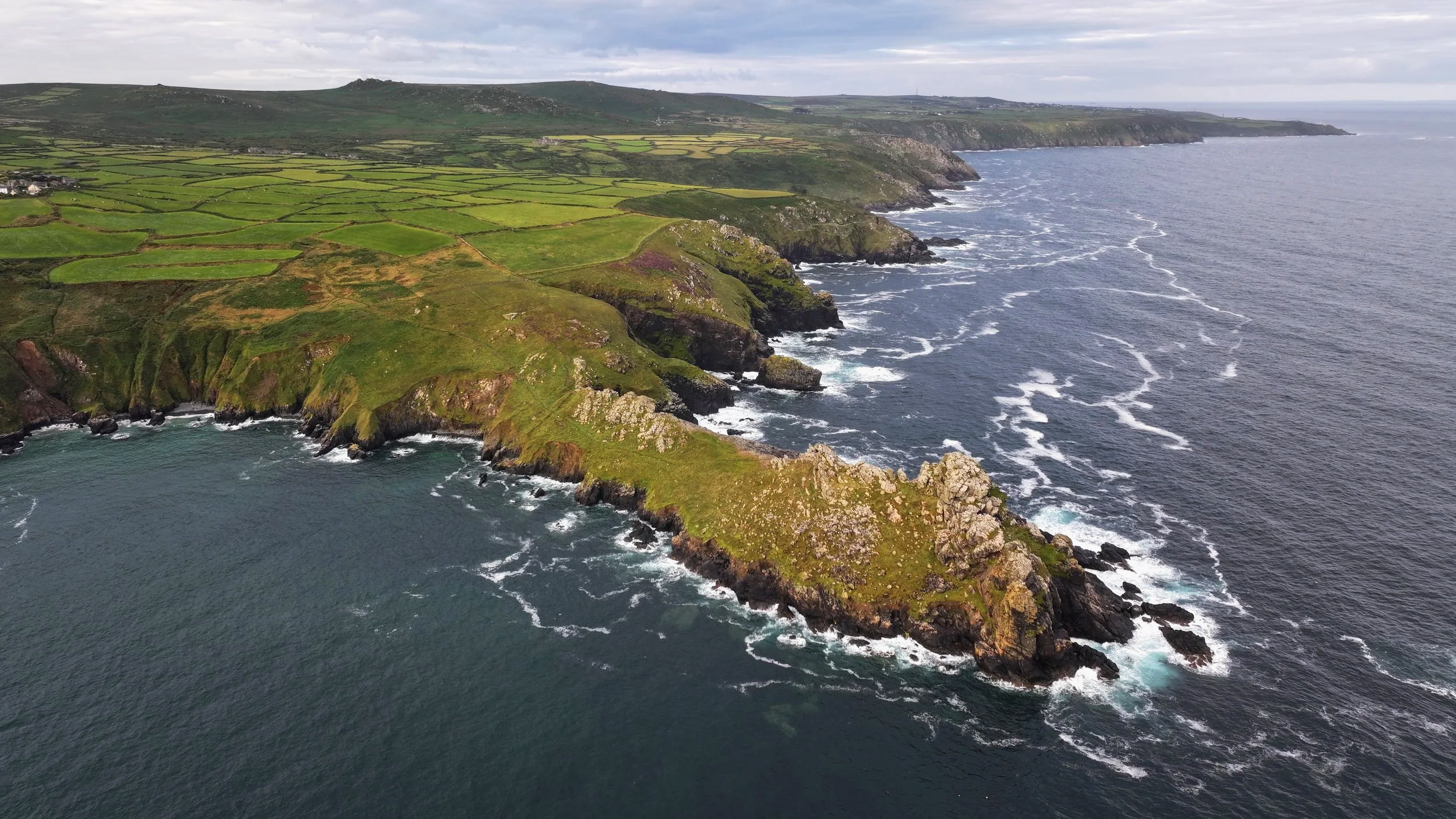 Aerial view of rugged coastline with green grass-covered cliffs extending into the ocean, with waves crashing against rocks at the base of the cliffs. The sea is partly cloudy with a gray sky.