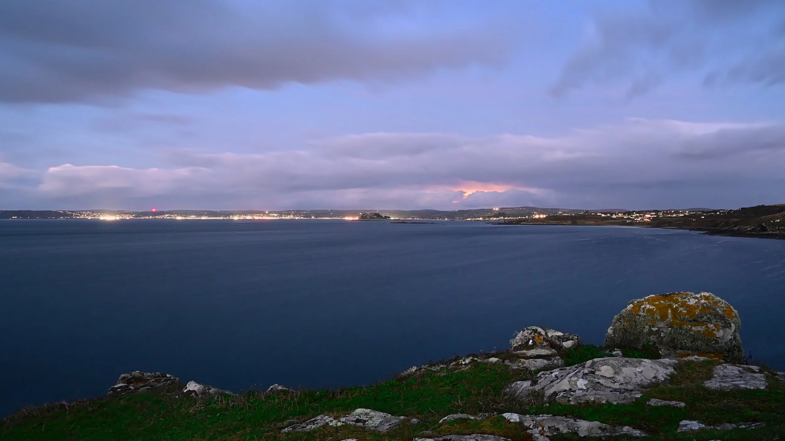 A coastal landscape during twilight, showing a calm sea in the foreground, rocky shoreline with patches of grass, and distant city lights along the horizon under a cloudy sky.