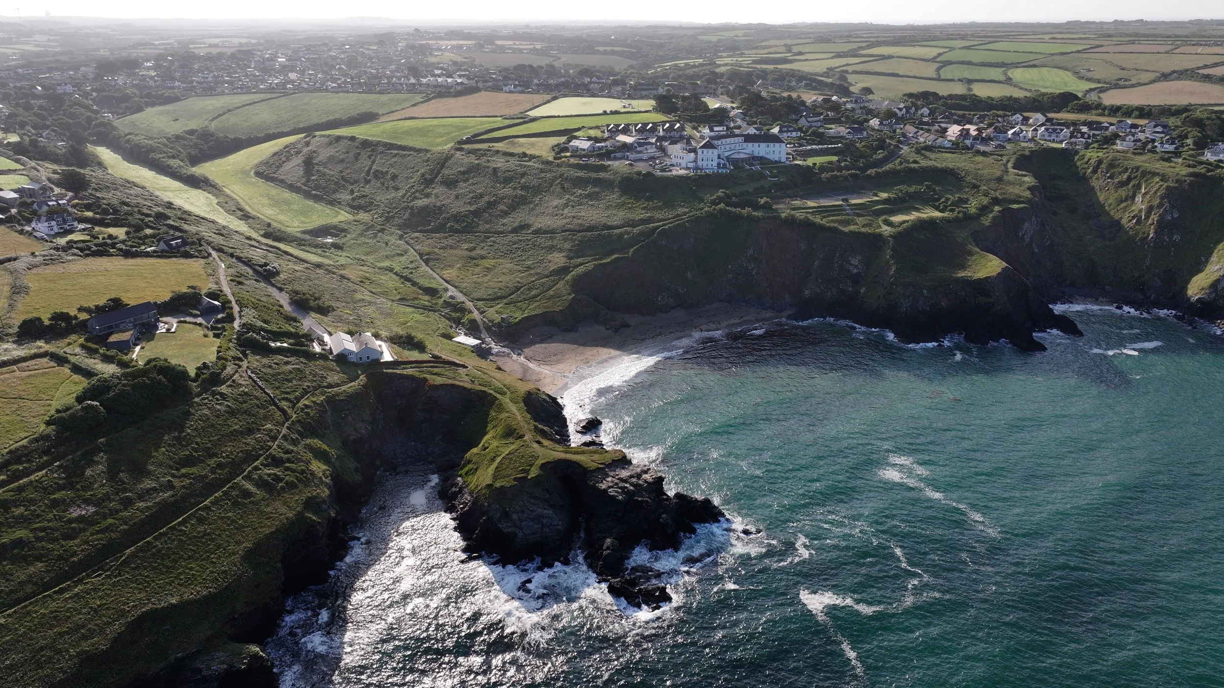 Aerial view of a coastal village with green hills, houses, and cliffs overlooking the ocean, with waves crashing against the rocks at the shore.