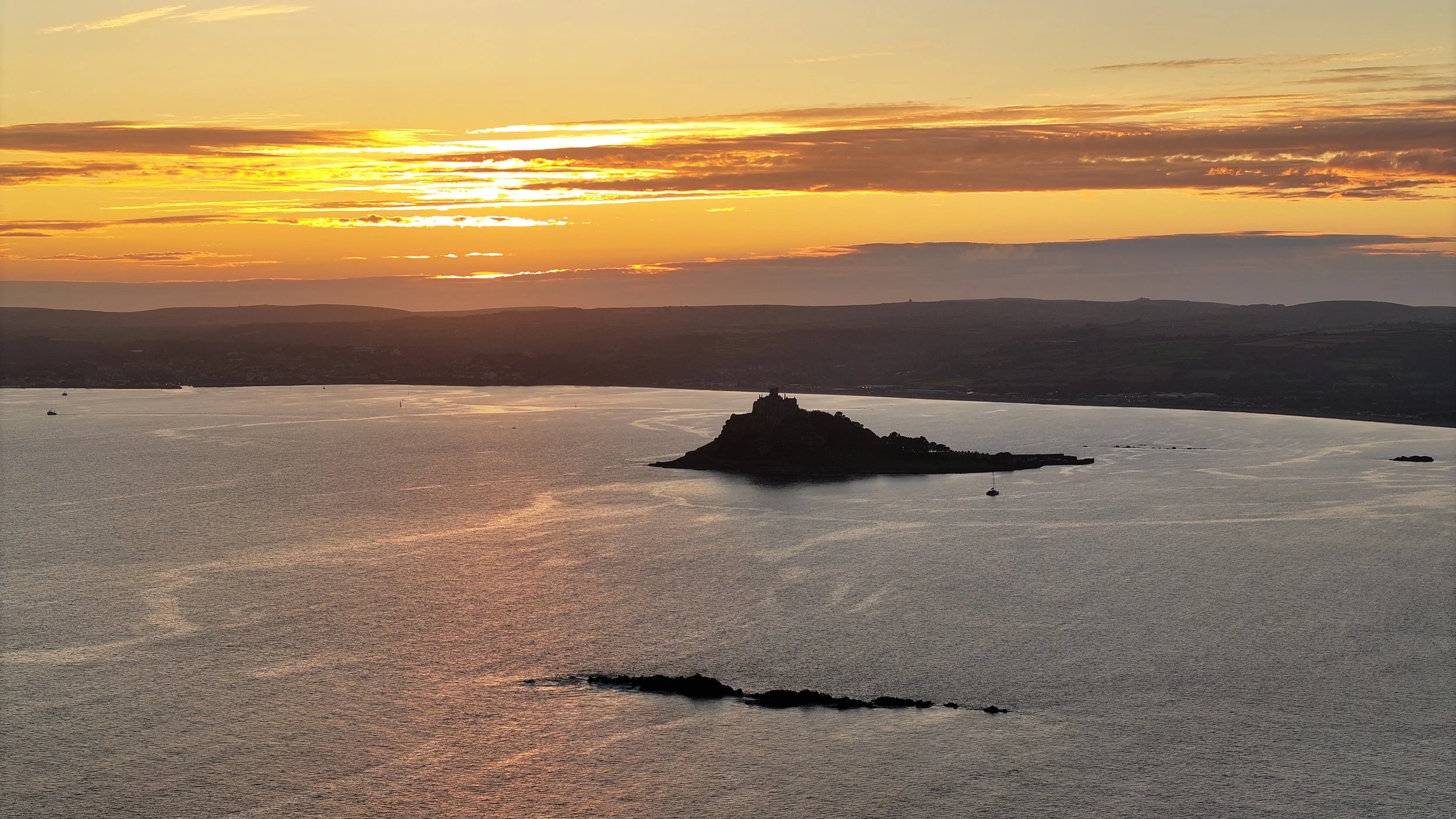 Aerial sunset view of St Michael’s Mount in Marazion, Cornwall, silhouetted against golden evening light over Mount’s Bay.