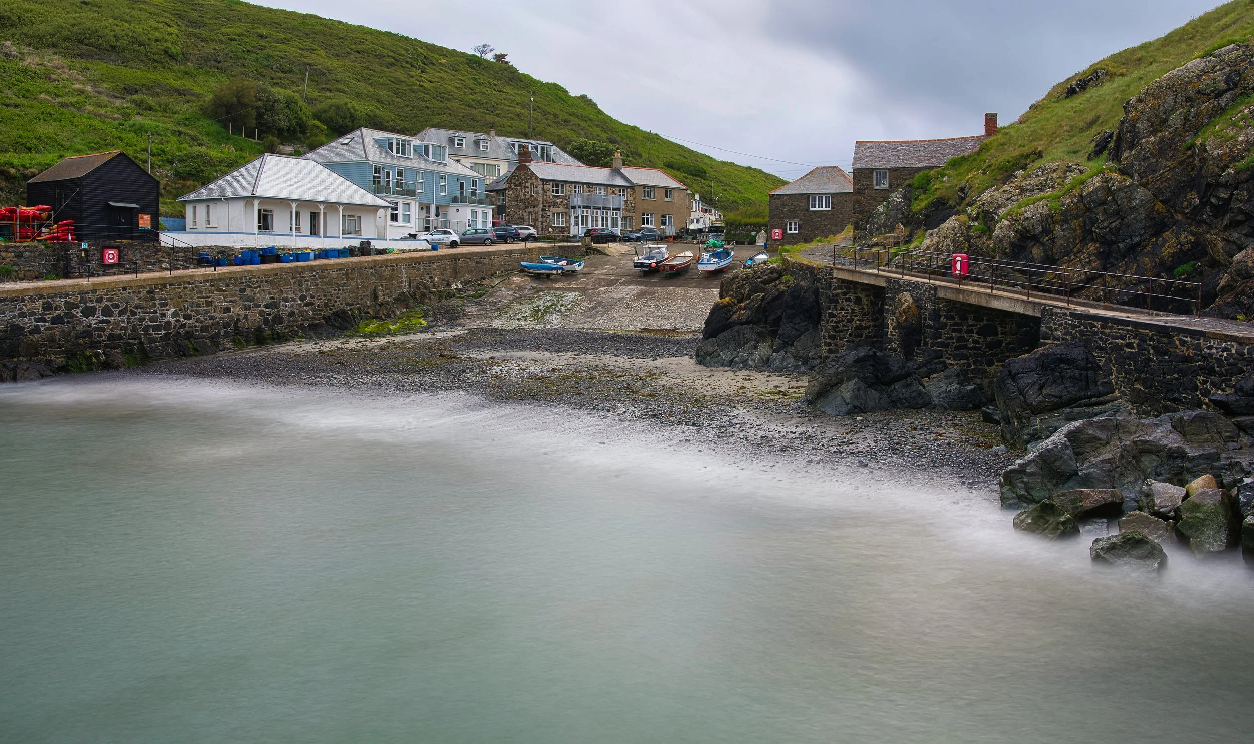 A small harbor with boats docked along a stone pier, in front of a row of houses on a hillside, under gray cloudy skies.