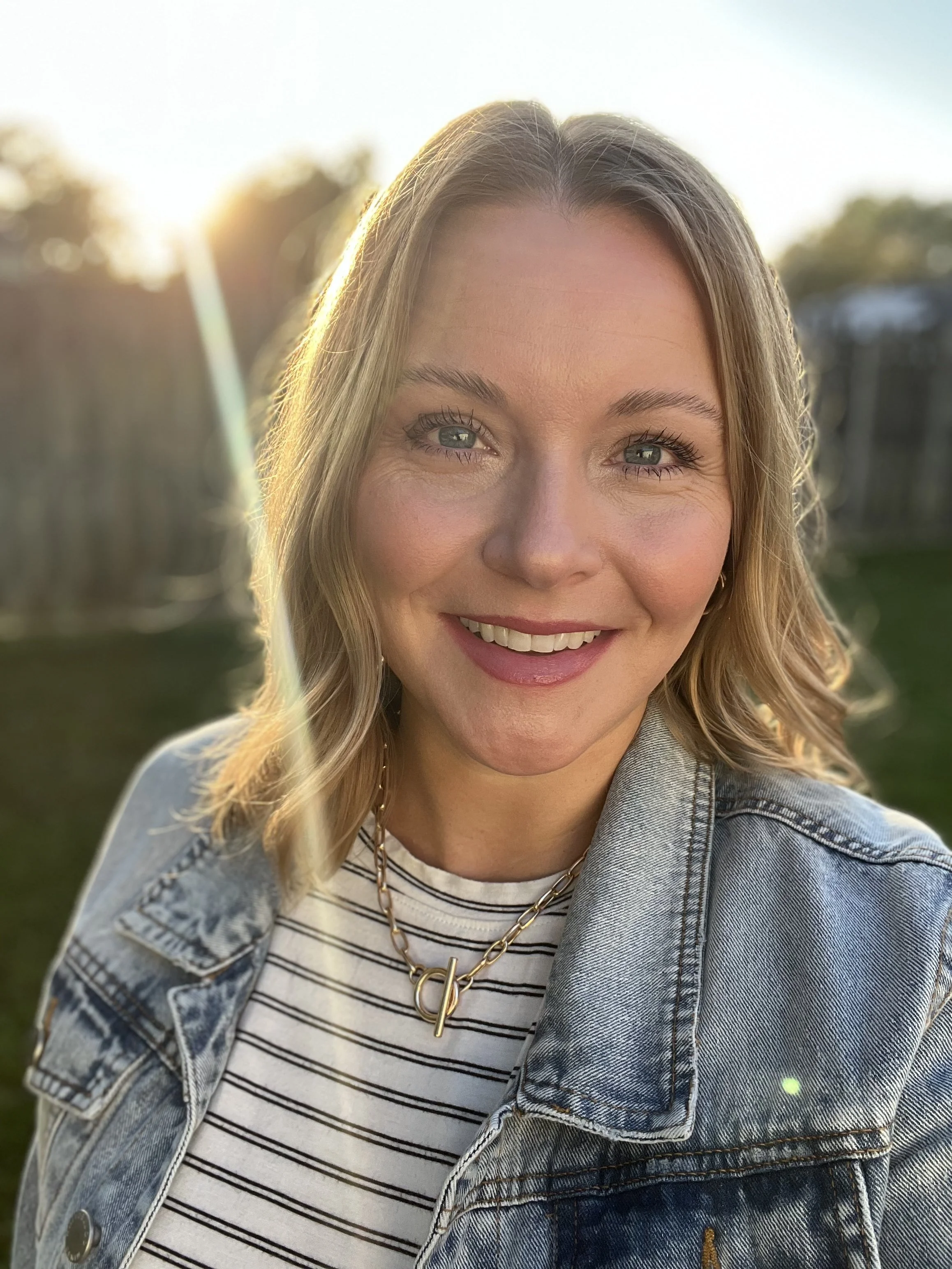 A smiling woman with blonde hair, blue eyes, wearing a striped shirt, denim jacket, and gold necklace, outdoors at sunset.
