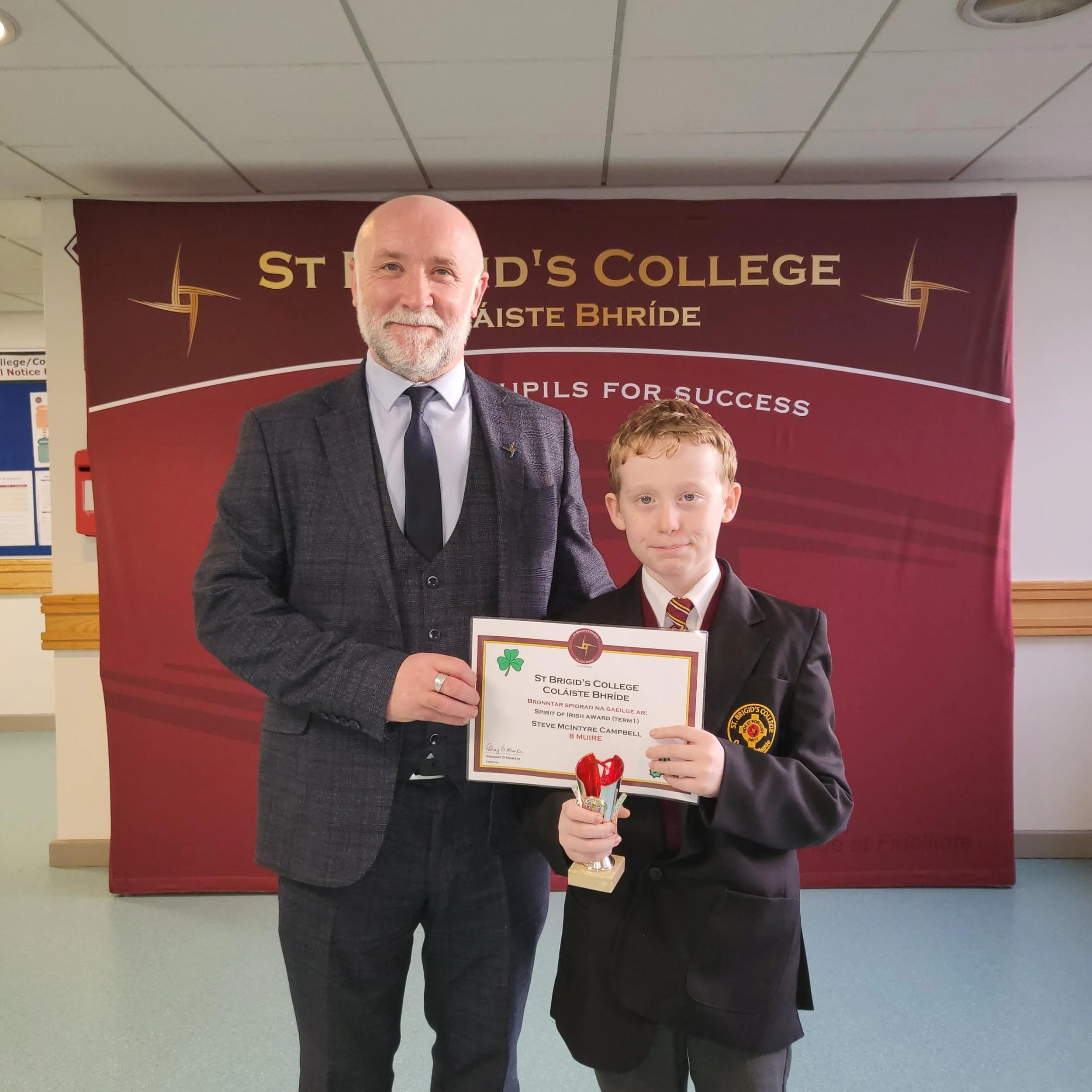 A man and a boy standing together in front of a red backdrop at St. Brigid's College, holding a certificate and a small trophy. The man has a beard and is wearing a gray suit with a tie, while the boy has red hair and is dressed in a black blazer wit