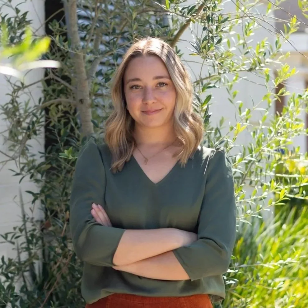 A young woman standing outdoors with her arms crossed, surrounded by greenery and sunlight, in front of a white brick wall.